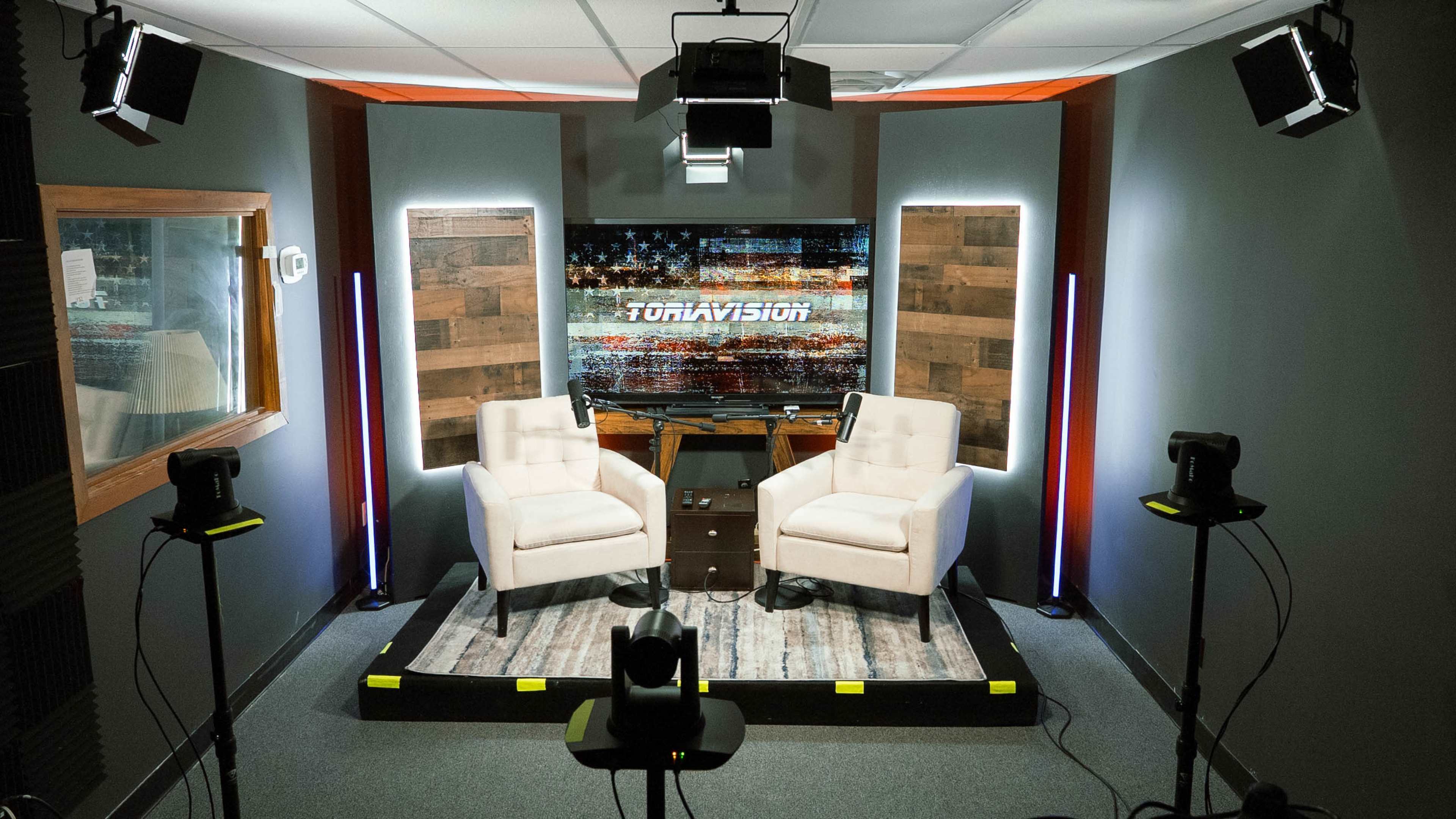 The image shows a television studio setup with two chairs, a small table, and various lighting equipment arranged around a wooden backdrop.