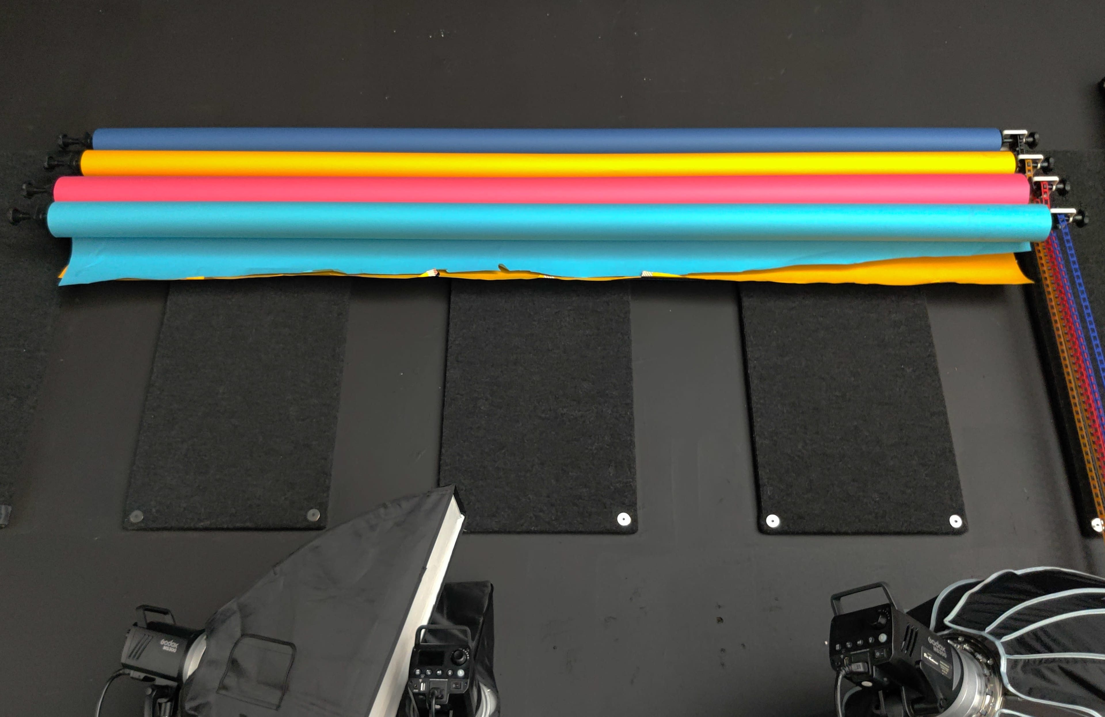 A photography studio setup with colorful backdrop rolls above dark mats and light equipment positioned on the floor.