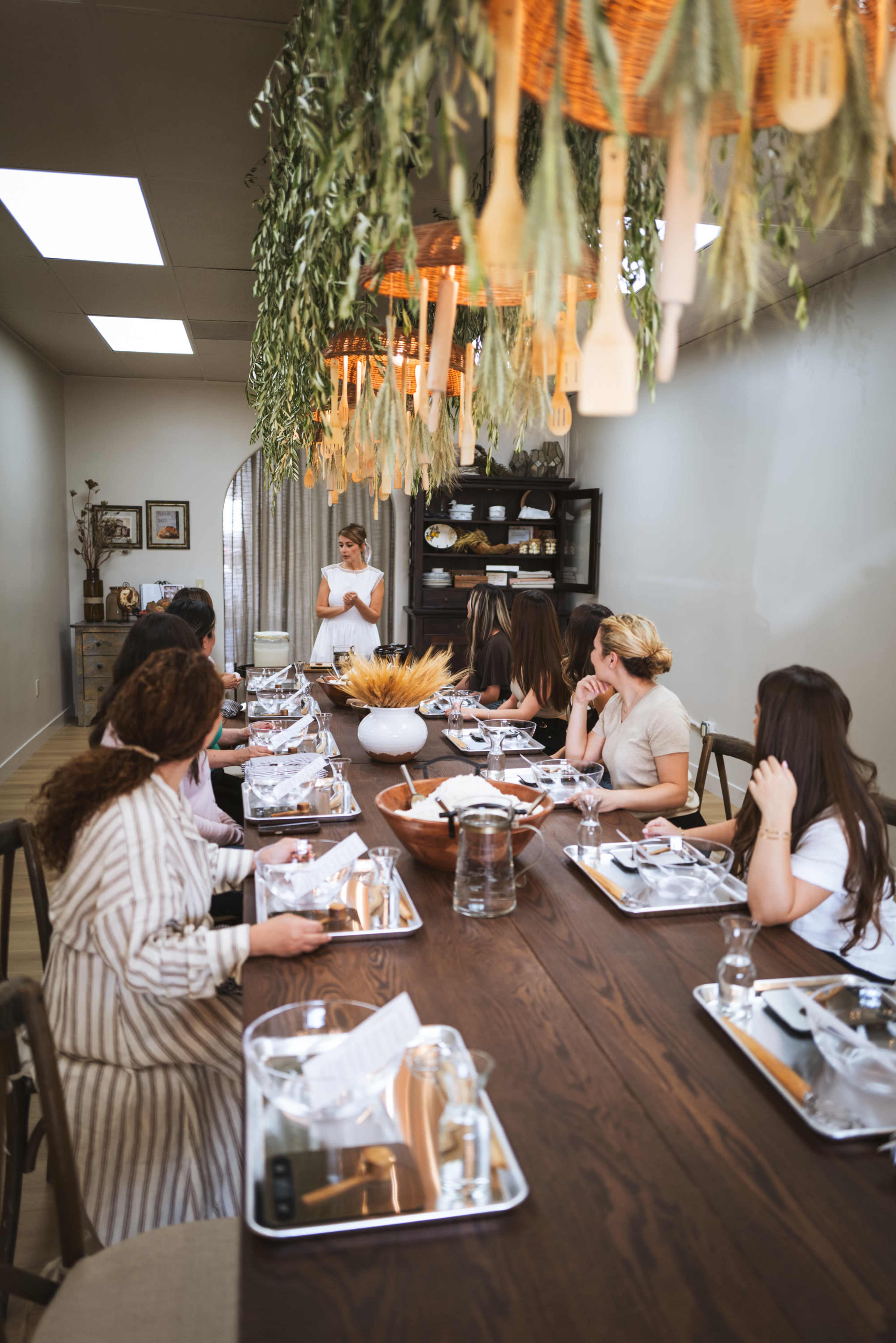 A long dining table is set for a meal, with seated guests listening to a speaker at the end of the table in a decorated room with hanging greenery.