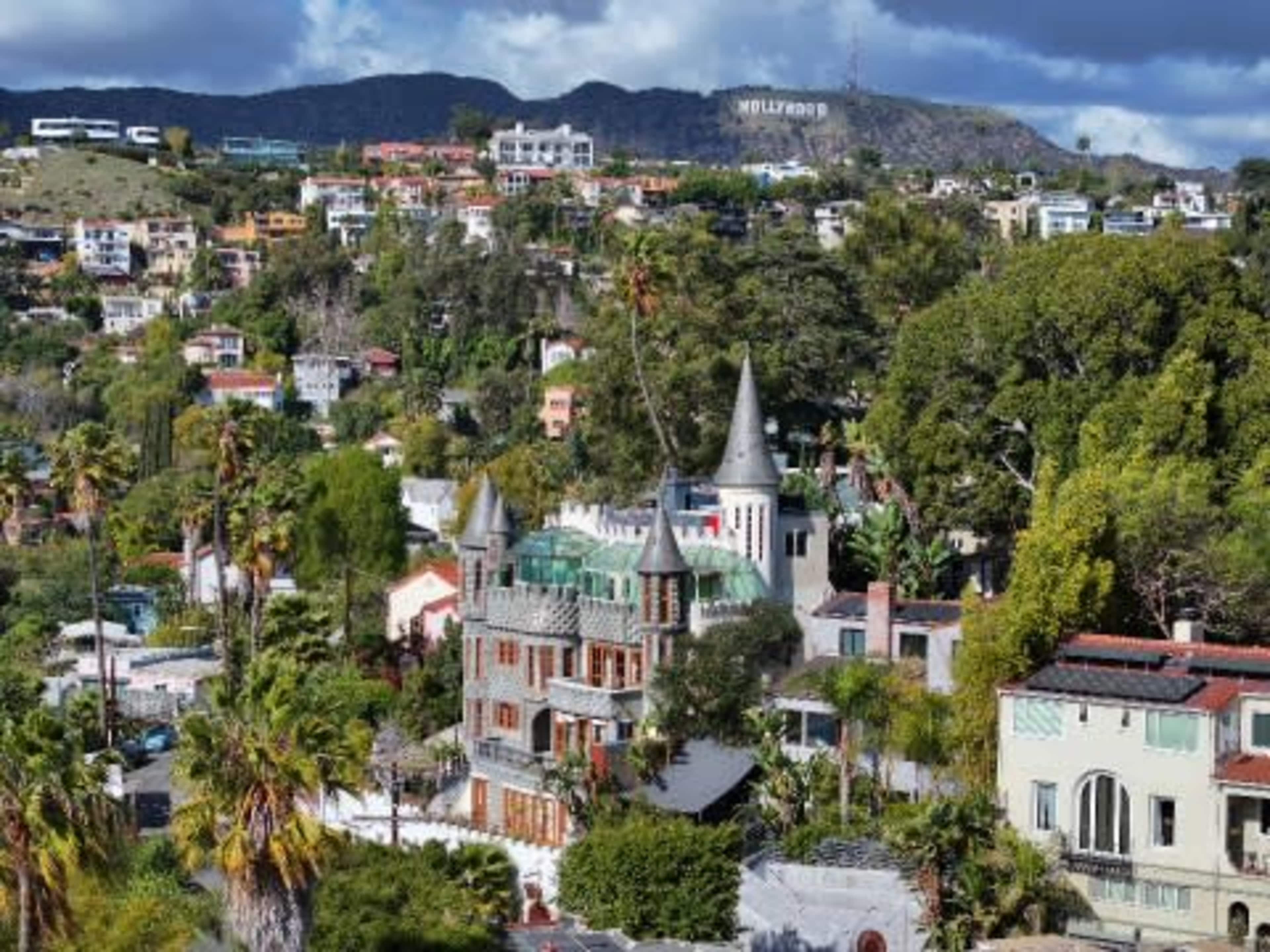 The image shows a hillside view of a neighborhood with various houses, including a castle-like structure, and the Hollywood sign visible in the background.