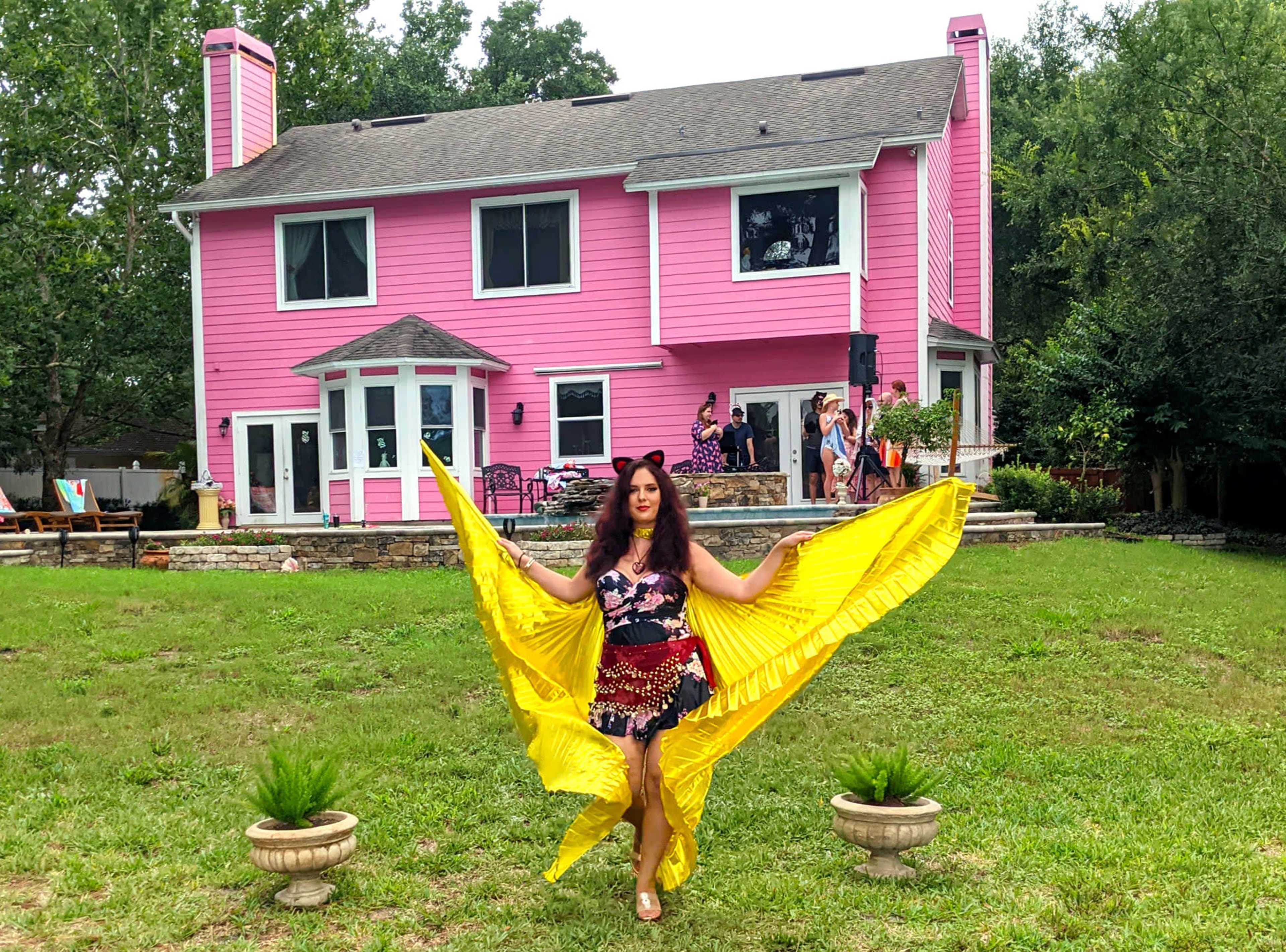 A woman in a colorful outfit holds a yellow fabric as she stands in front of a large pink house surrounded by greenery.
