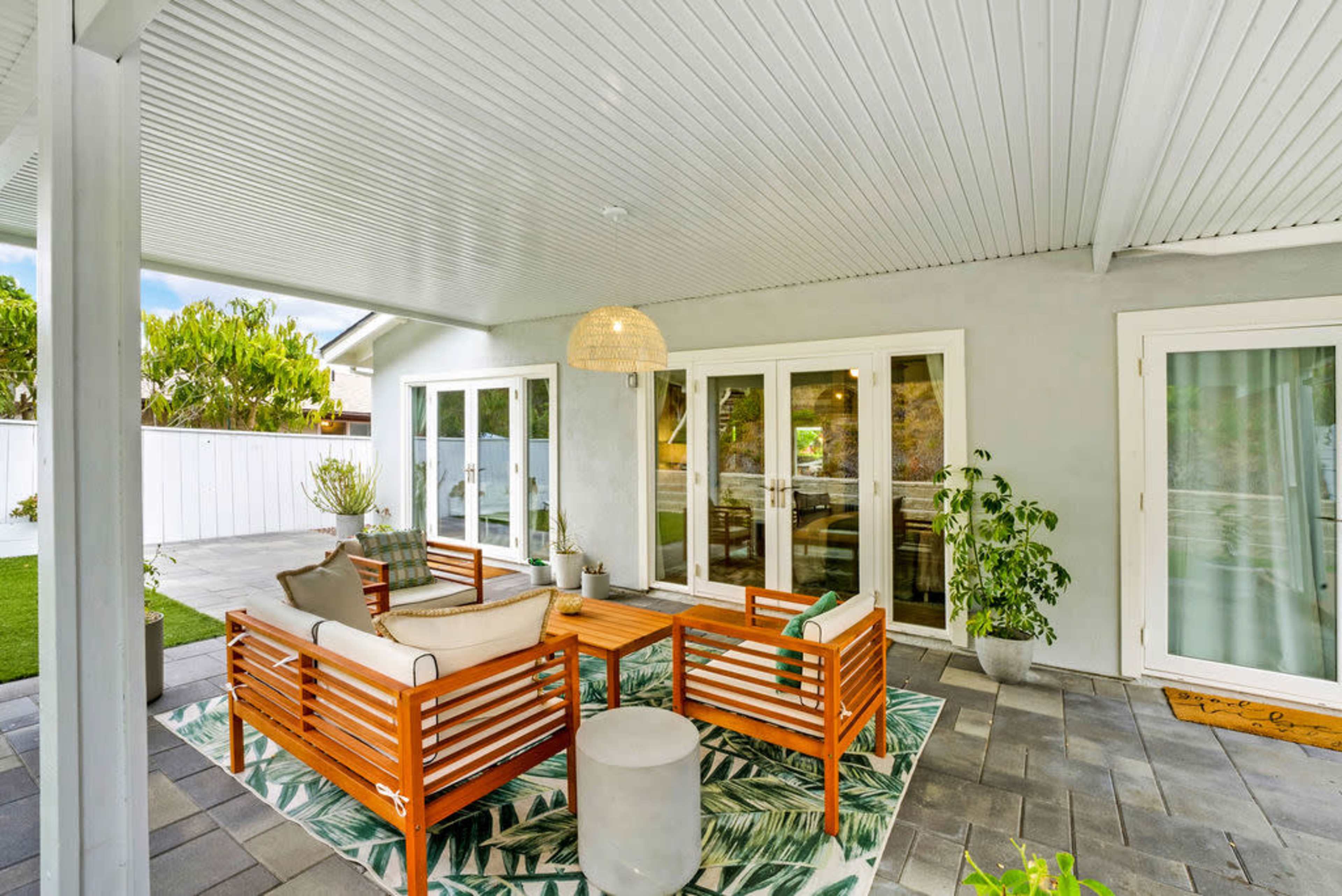 A covered patio features wooden furniture arranged around a central table, with potted plants and a modern light fixture hanging above.