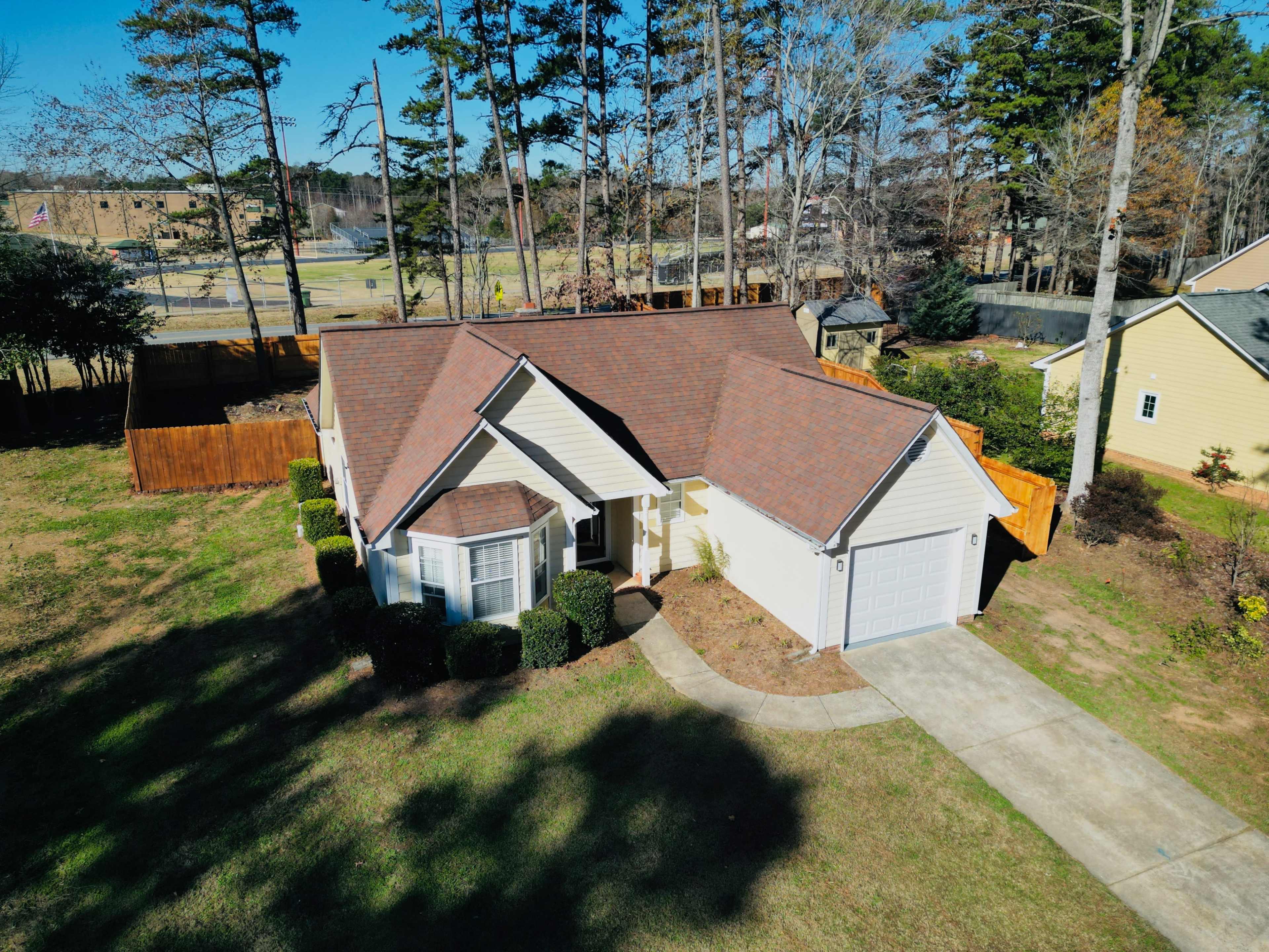A single-story house with a brown roof and a neatly landscaped yard is located in a suburban neighborhood, surrounded by trees and a wooden fence.