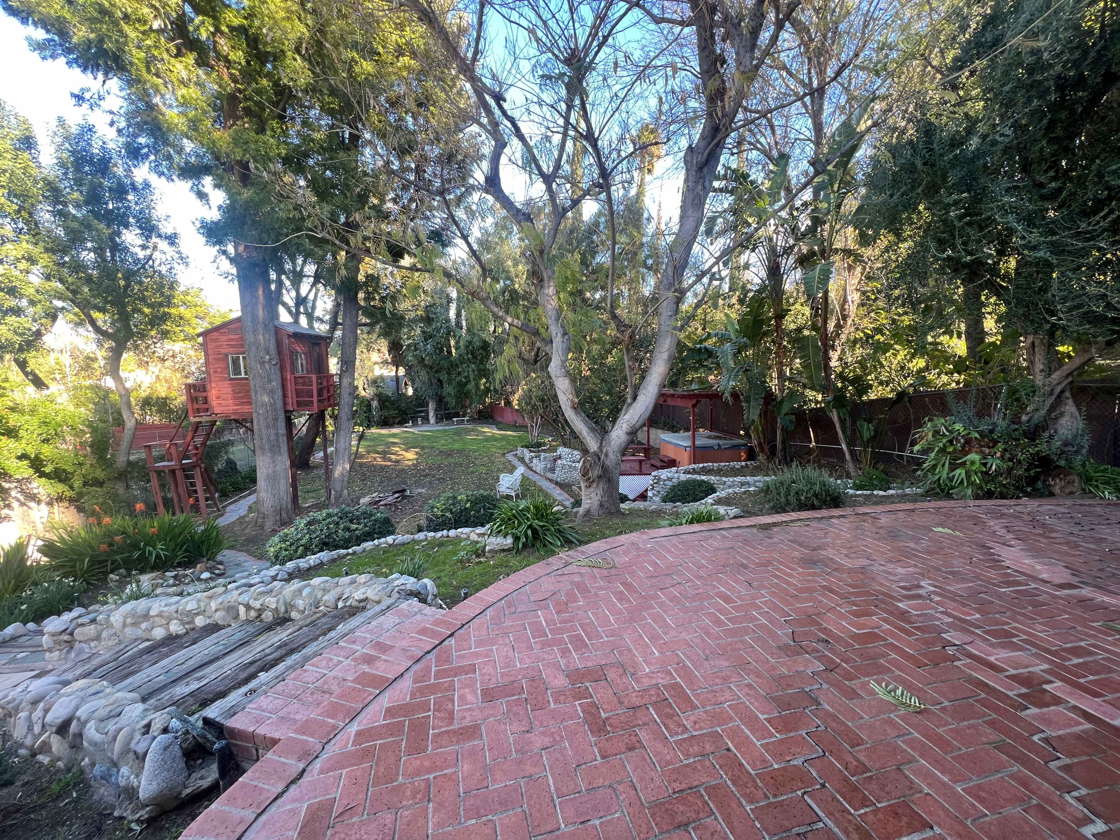 A landscaped yard featuring a brick pathway, a large tree, a wooden playhouse, and a gazebo surrounded by greenery.