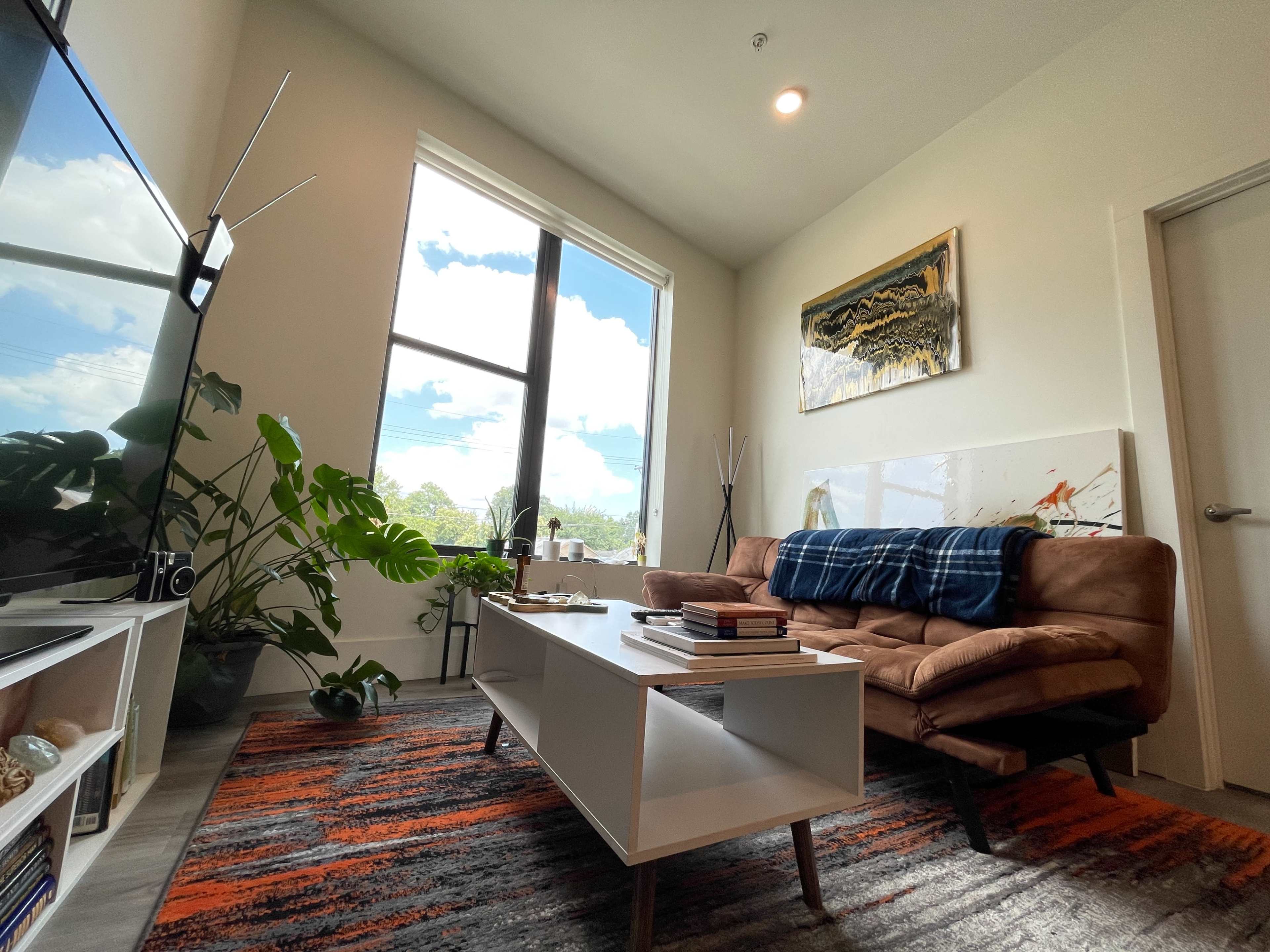 A modern living room with a brown sofa, a coffee table stacked with books, a large window allowing natural light, and indoor plants.