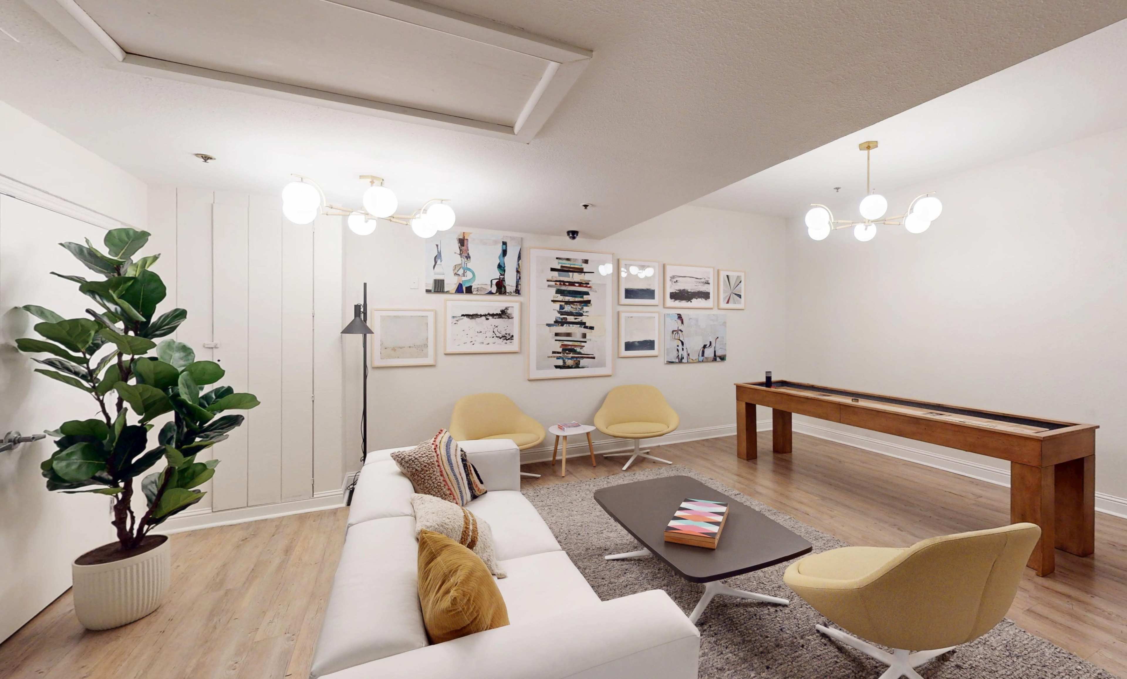 A modern lounge area features a white sofa, two yellow chairs, and a wooden table, with artwork displayed on the walls and a potted plant in one corner.