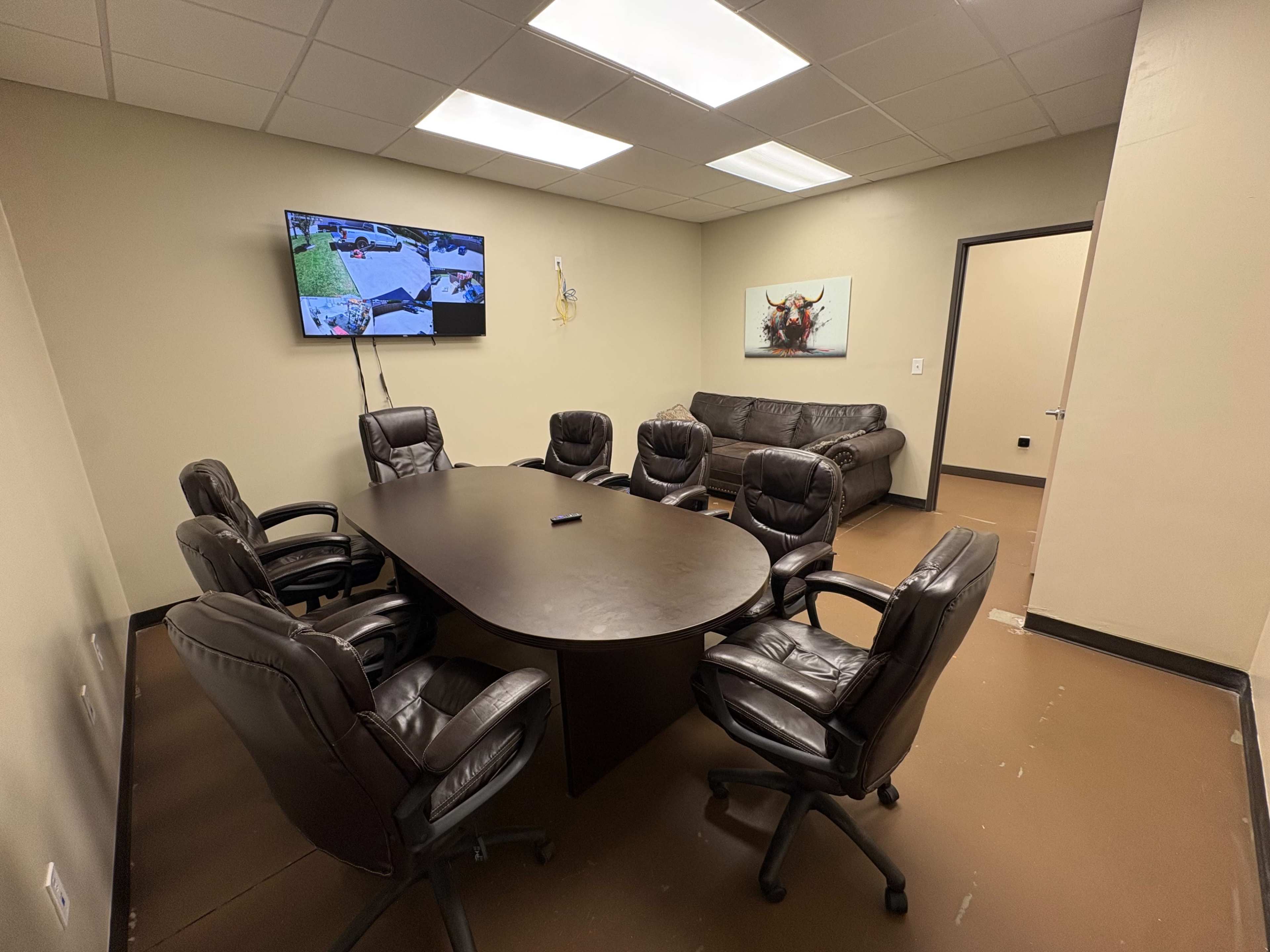 The image shows a conference room featuring a large oval table surrounded by six black leather chairs, a couch against one wall, and a television mounted on another wall.