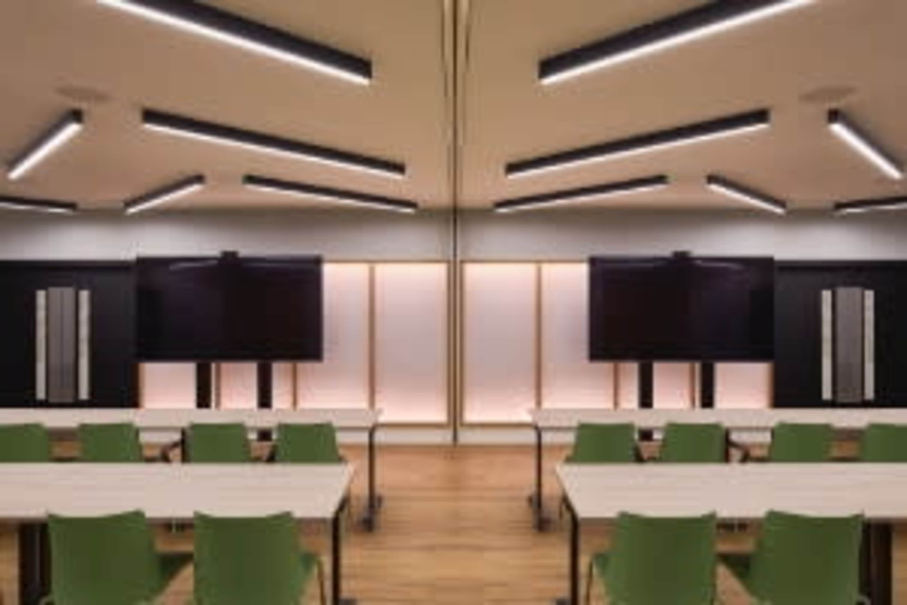 A modern classroom features rows of tables and chairs with two large screens mounted on the walls.