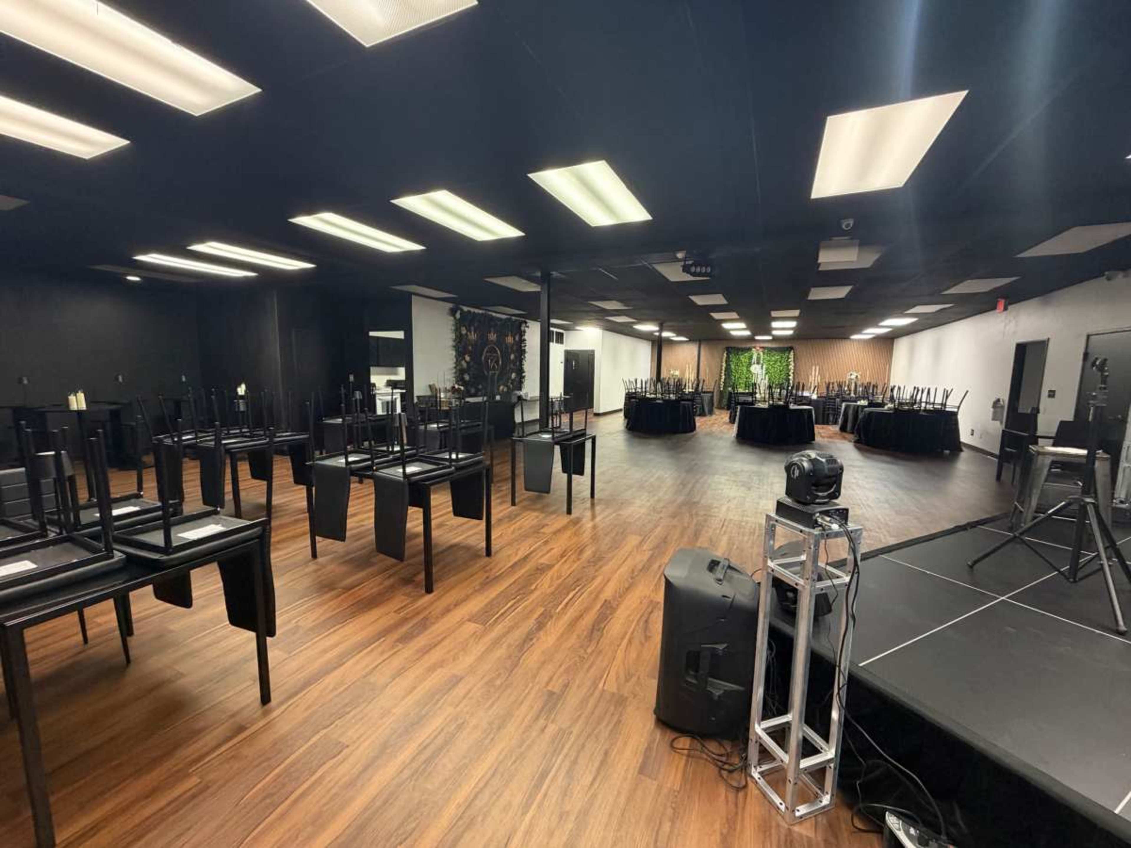 The image shows a large event space with black chairs stacked upside down on tables, while other tables are set up with black tablecloths in the background.