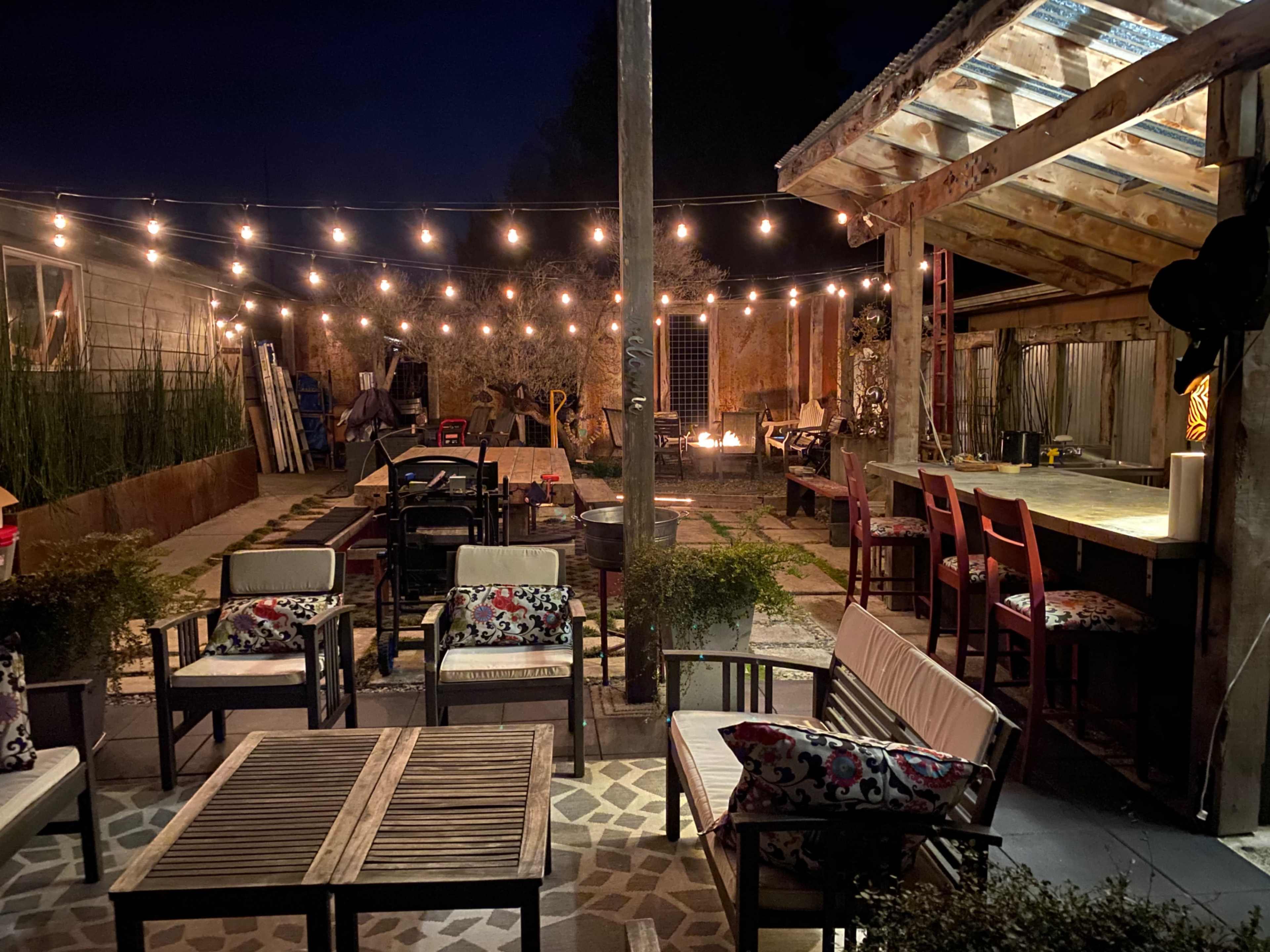 A nighttime outdoor patio area with string lights overhead, wooden seating, and a fire pit in the corner.
