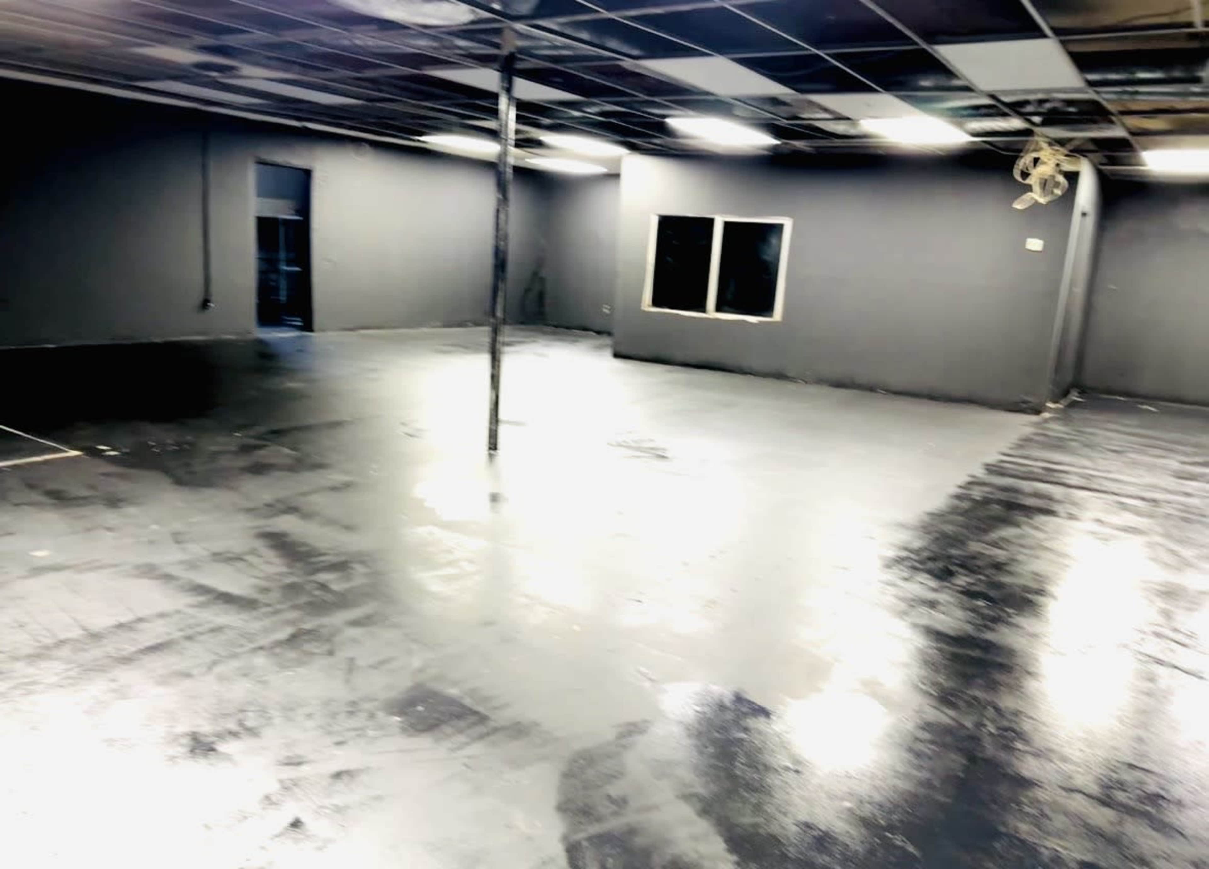 The image shows a sparsely furnished room with a glossy black floor, gray walls, and a few windows allowing light to enter.