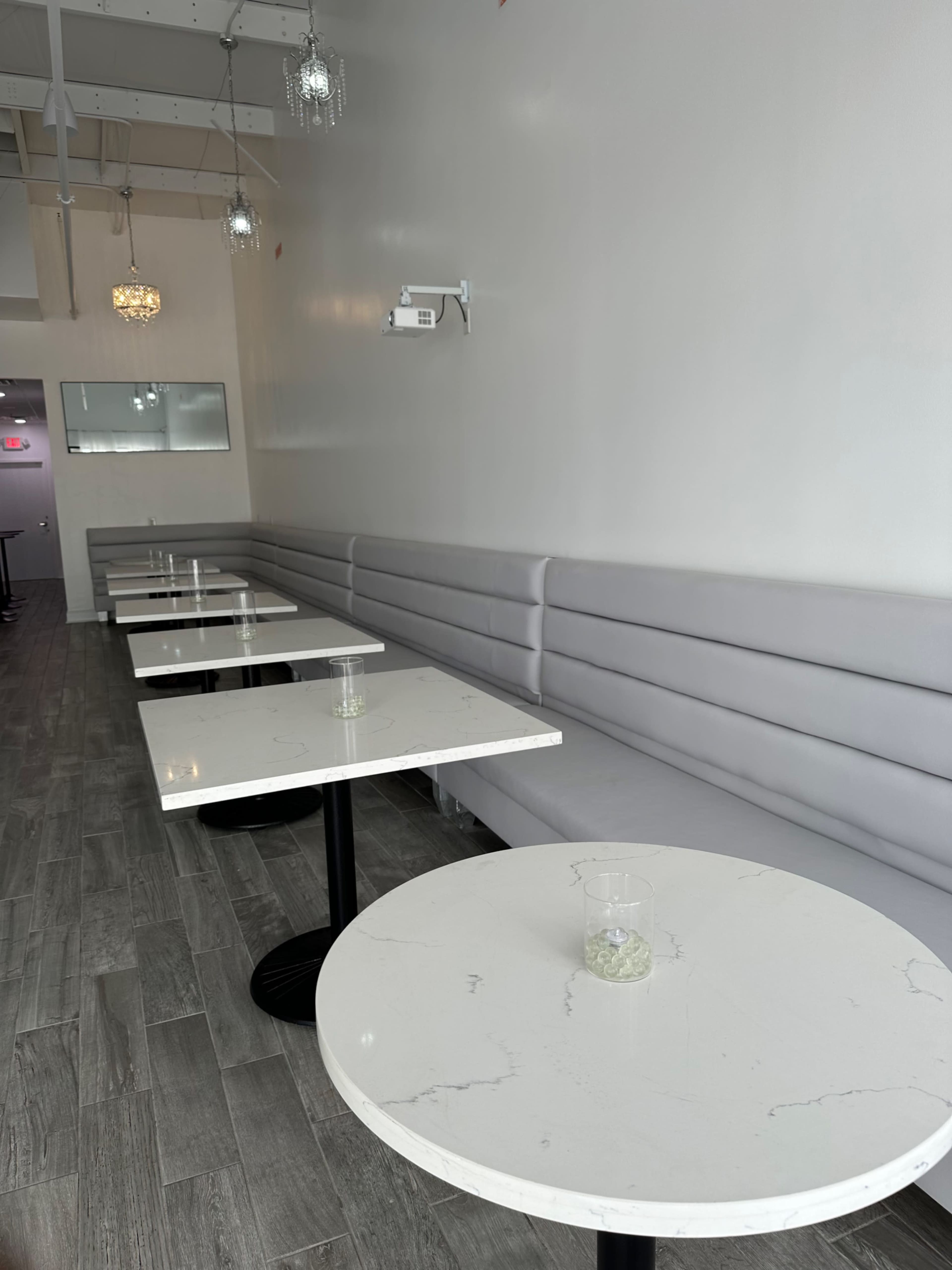 A minimalist cafe interior featuring several round tables with light-colored marble tops and a gray upholstered bench along a white wall.