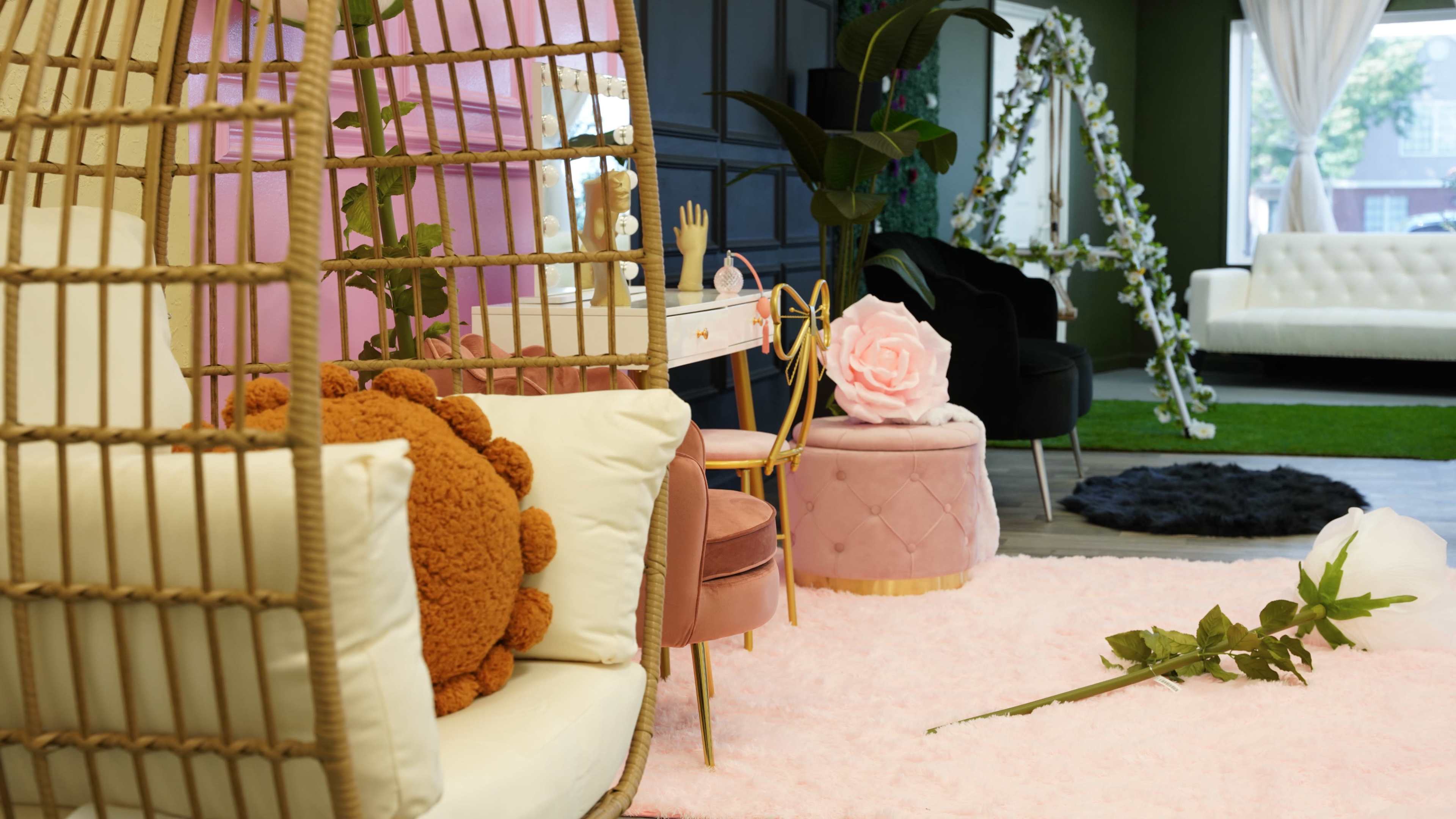 The scene features a stylish interior with a cozy seating area, including a rattan hanging chair, plush cushions, and decorative plants against a backdrop of colorful walls and furniture.