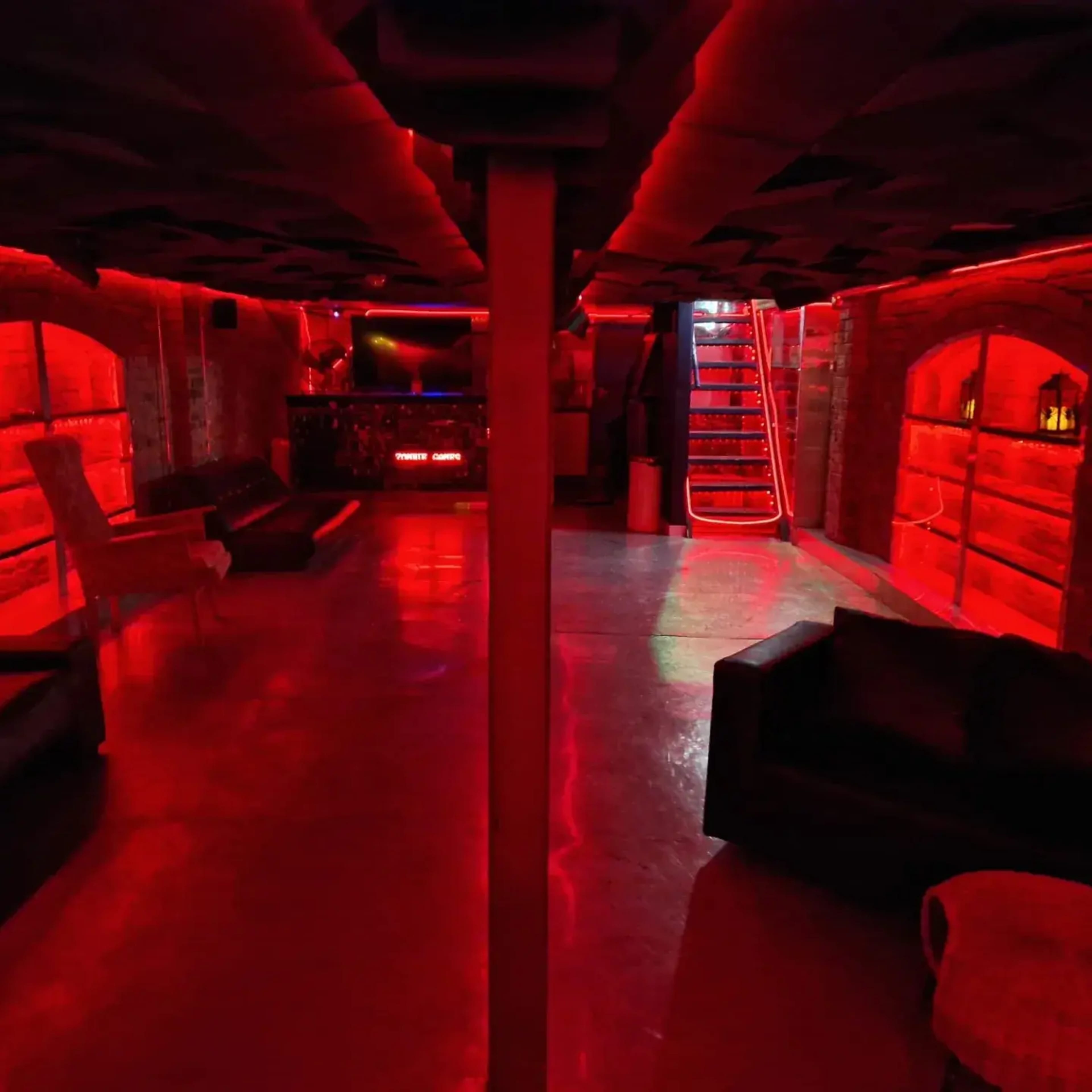 The image shows a dimly lit underground bar with red lighting, featuring a seating area and a bar counter in the background.