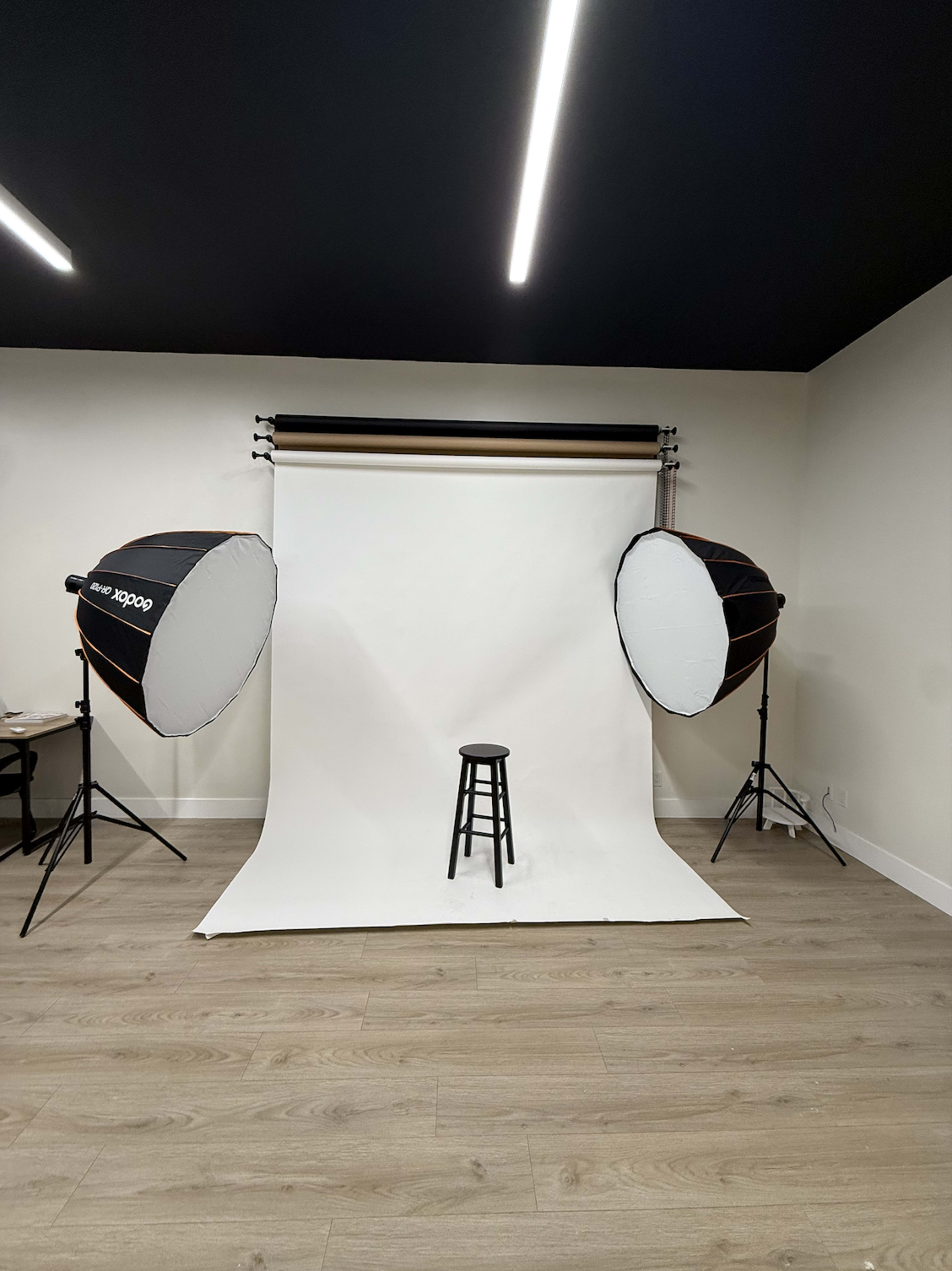 A photography studio features a white backdrop with a black stool positioned in front of two large softbox lights.