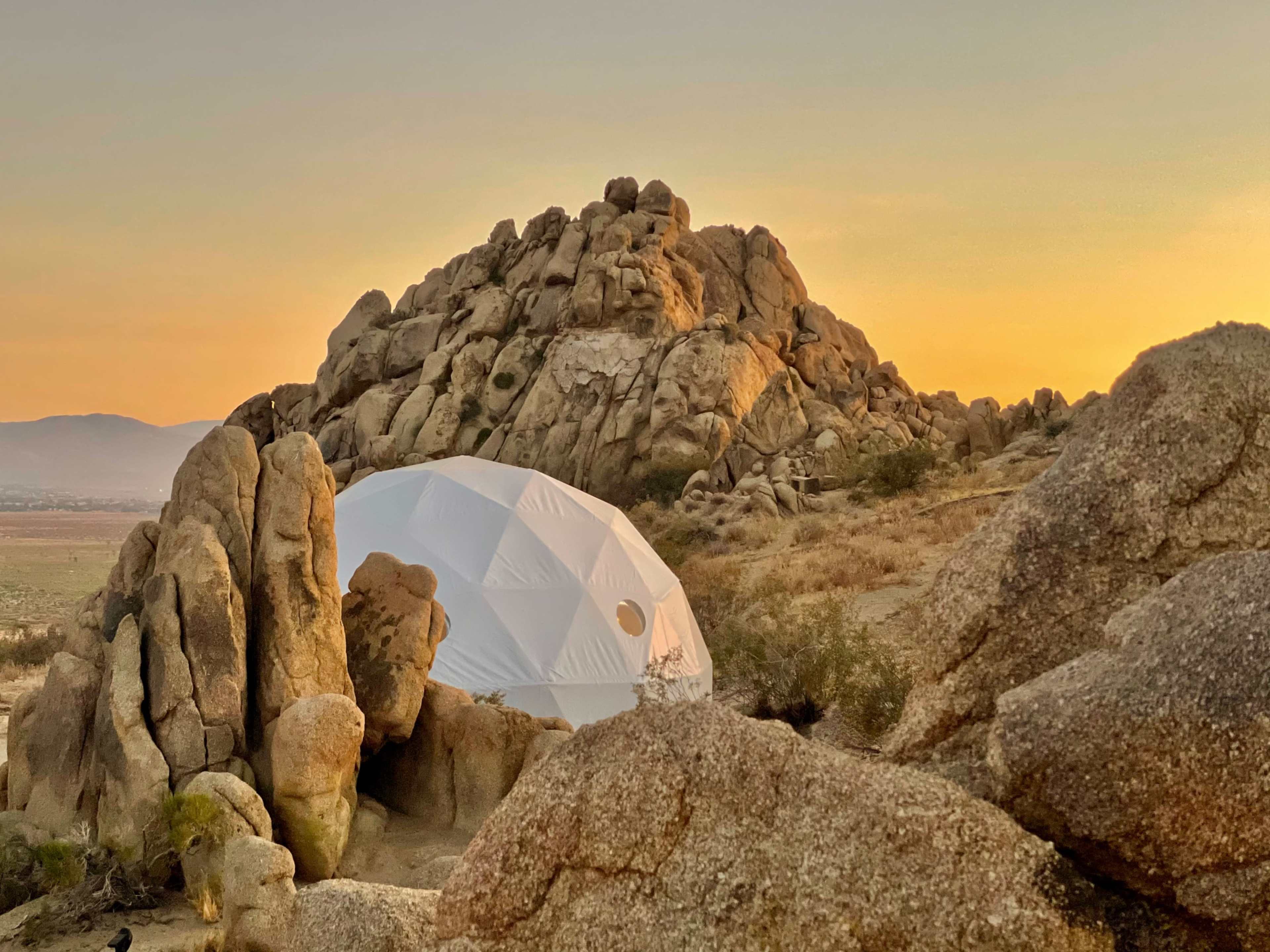 A white geodesic dome is nestled among large rock formations at sunset.