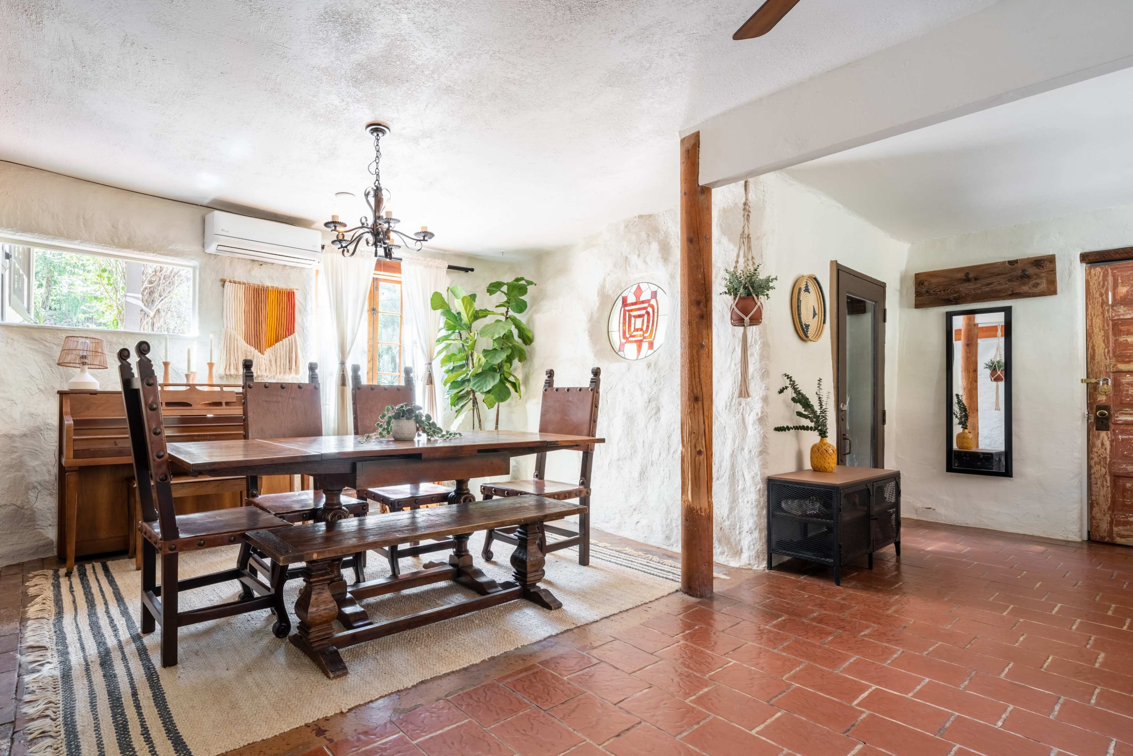 A rustic dining area featuring a large wooden table with benches, a chandelier, and decorative plants against textured white walls.