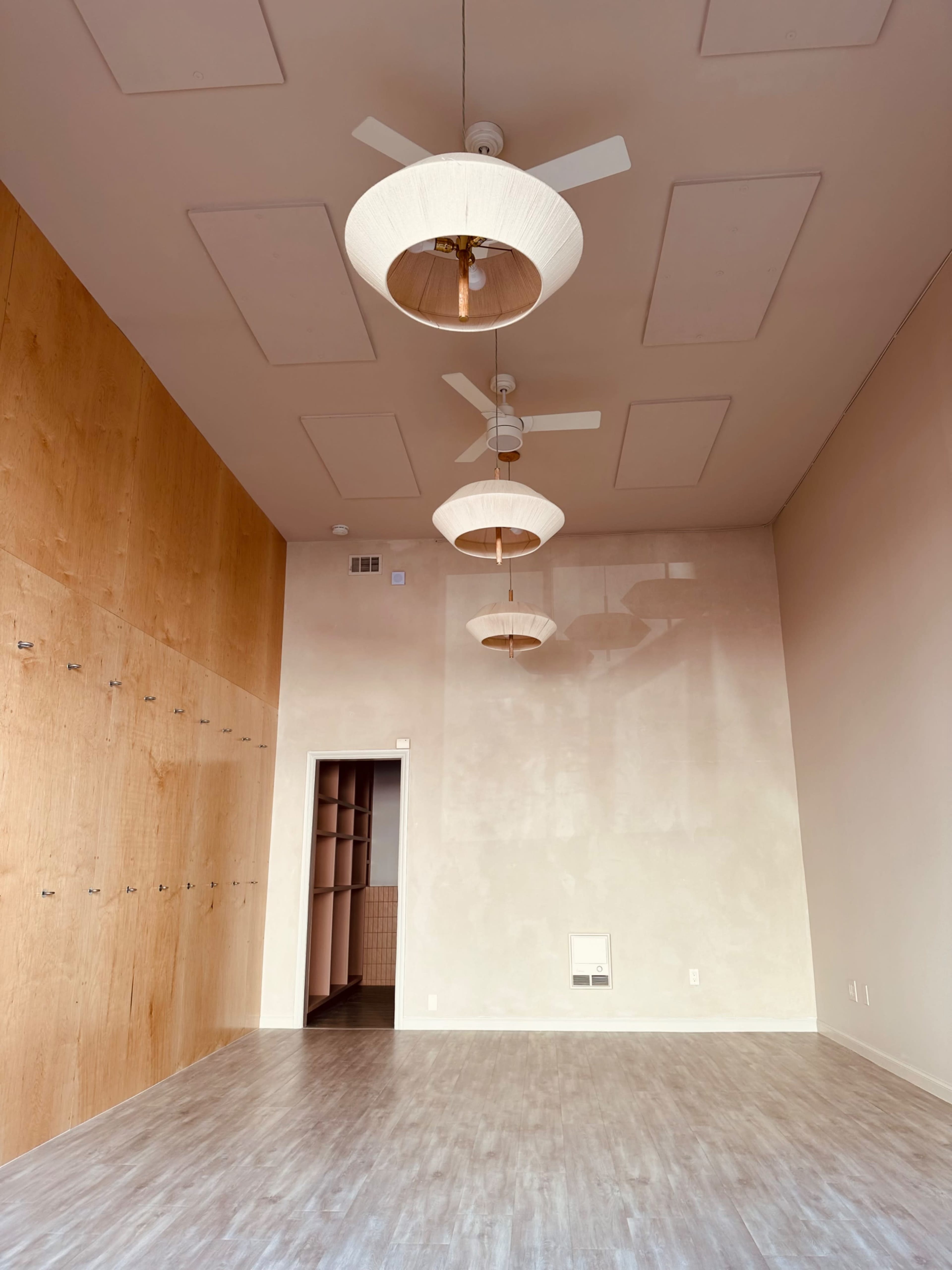 The image shows a vacant room with wooden paneling on one wall, three ceiling fans with light fixtures, and an adjoining door that leads to another area.