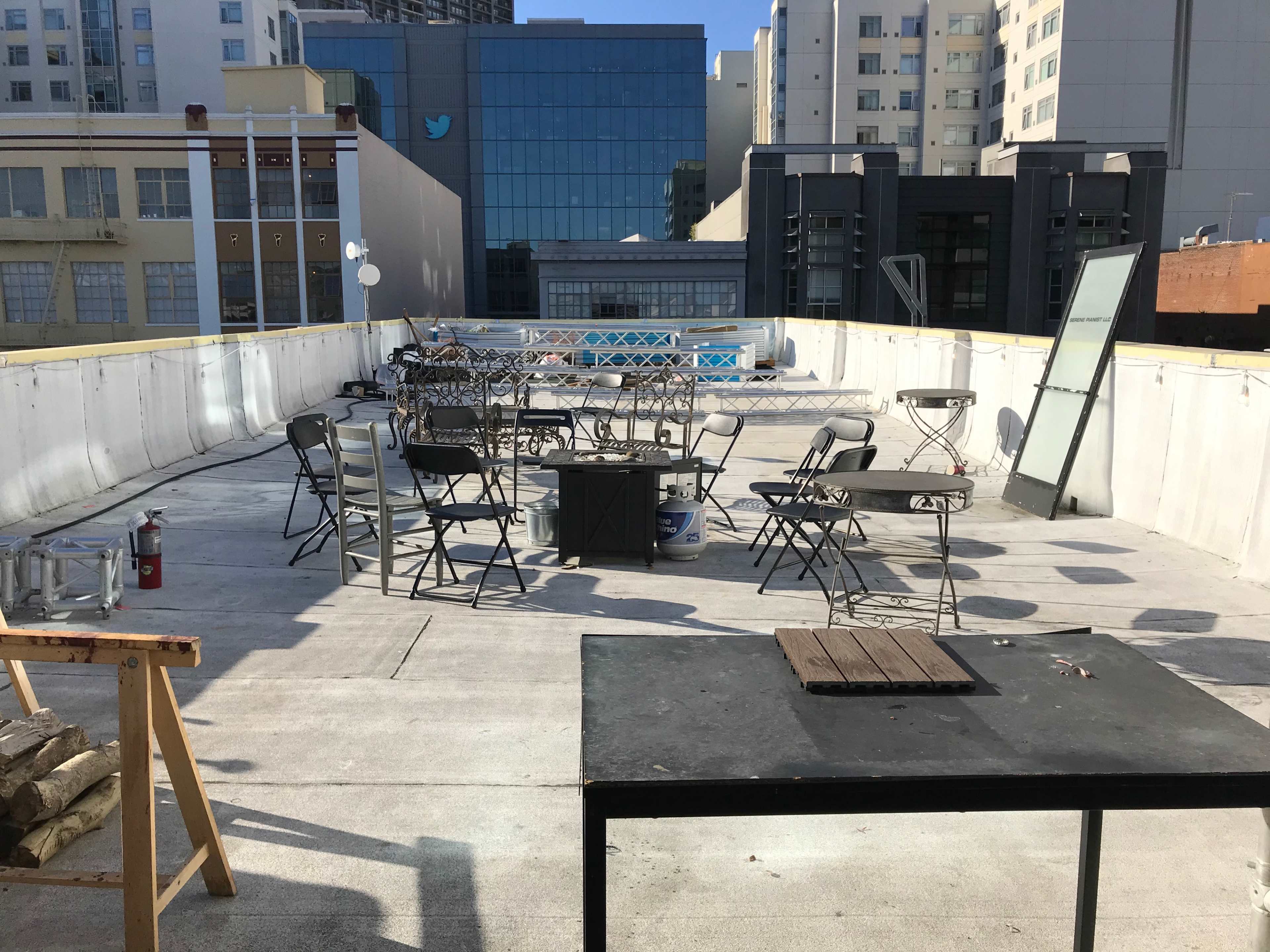 A rooftop space furnished with several empty tables and chairs, with buildings in the background.
