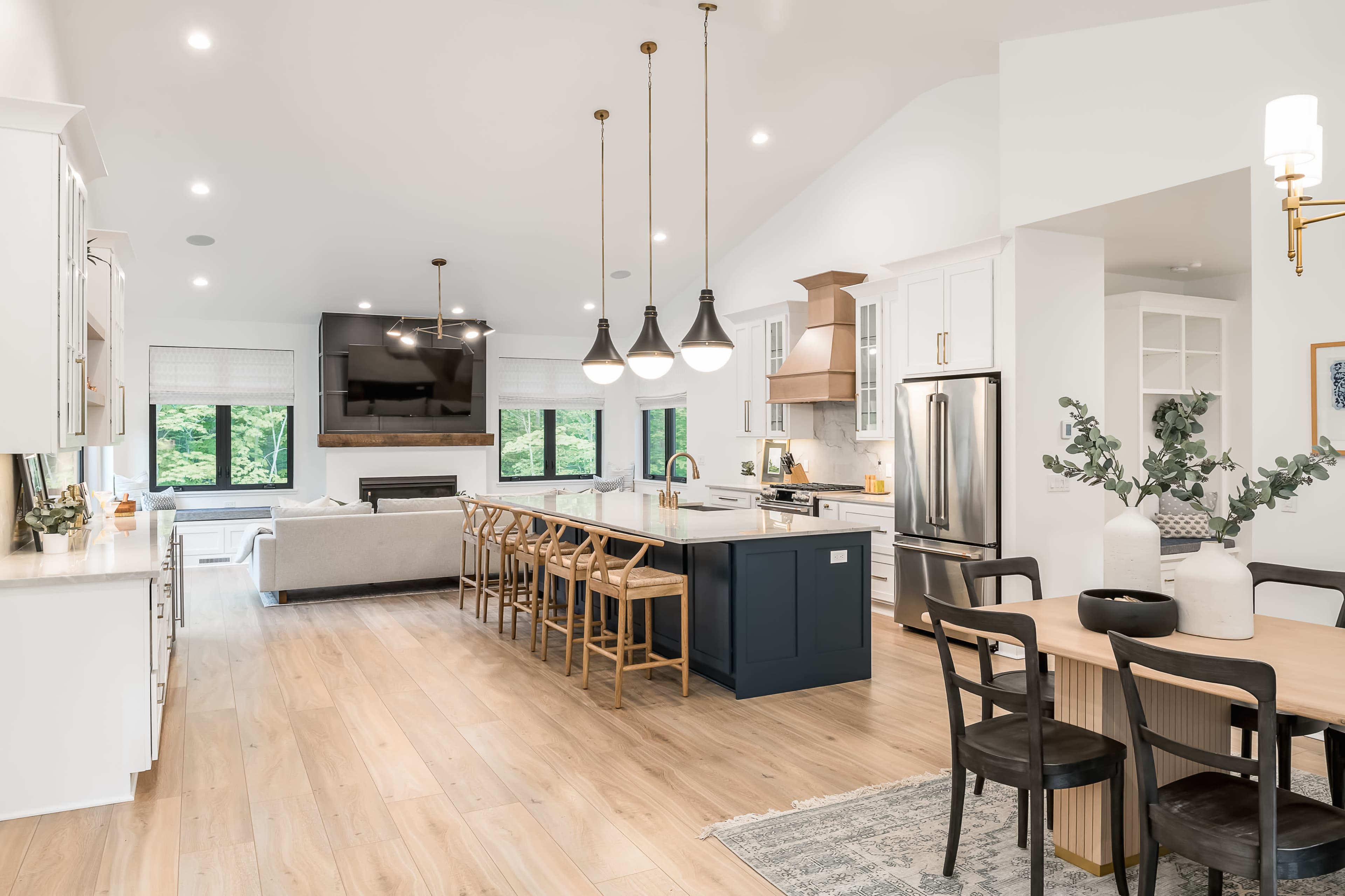 A modern kitchen and dining area featuring an island with bar stools, a dining table, and large windows overlooking a green outdoor space.
