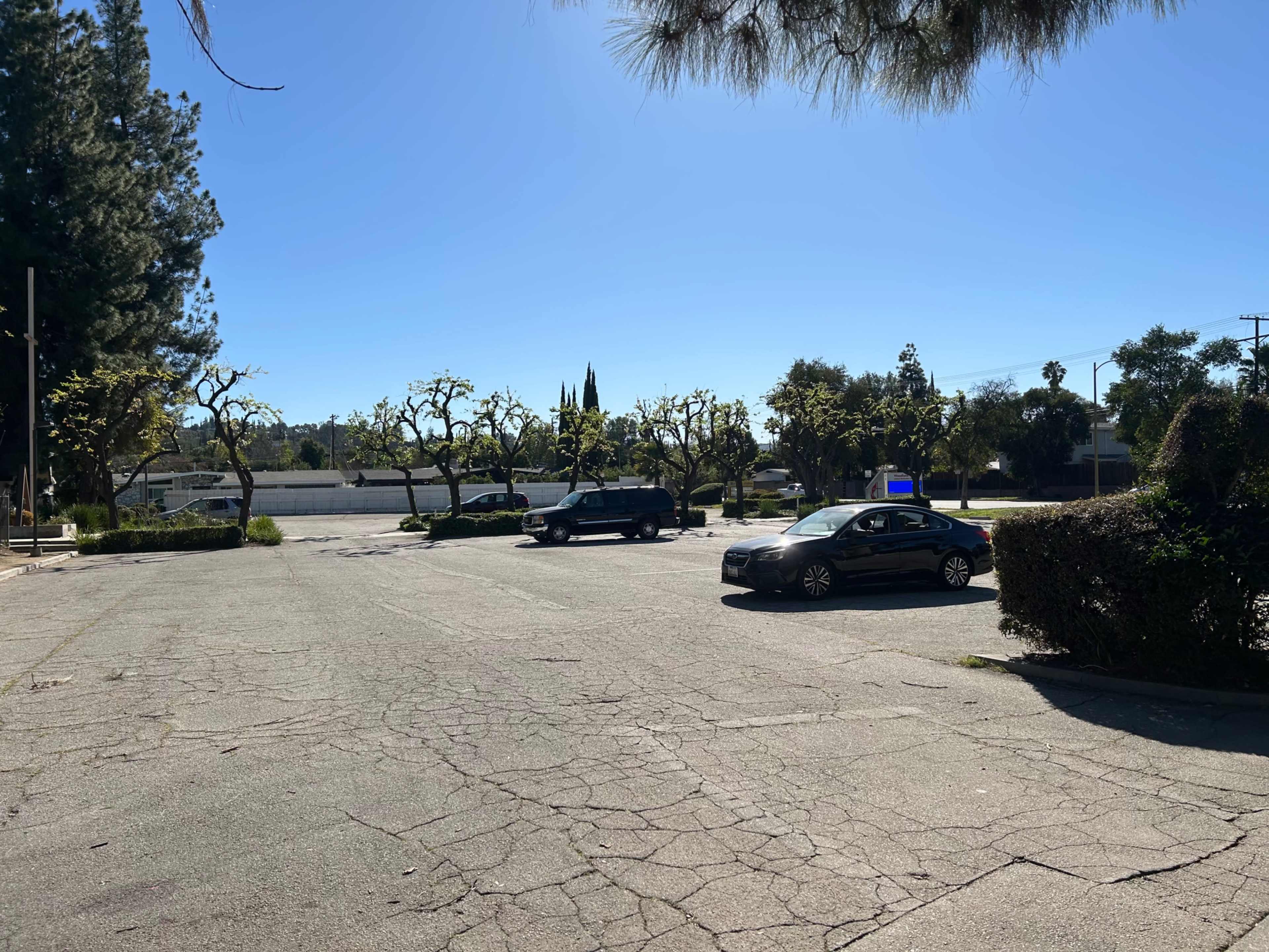 The image shows an empty parking lot with two cars parked on cracked pavement, surrounded by trees under a clear blue sky.