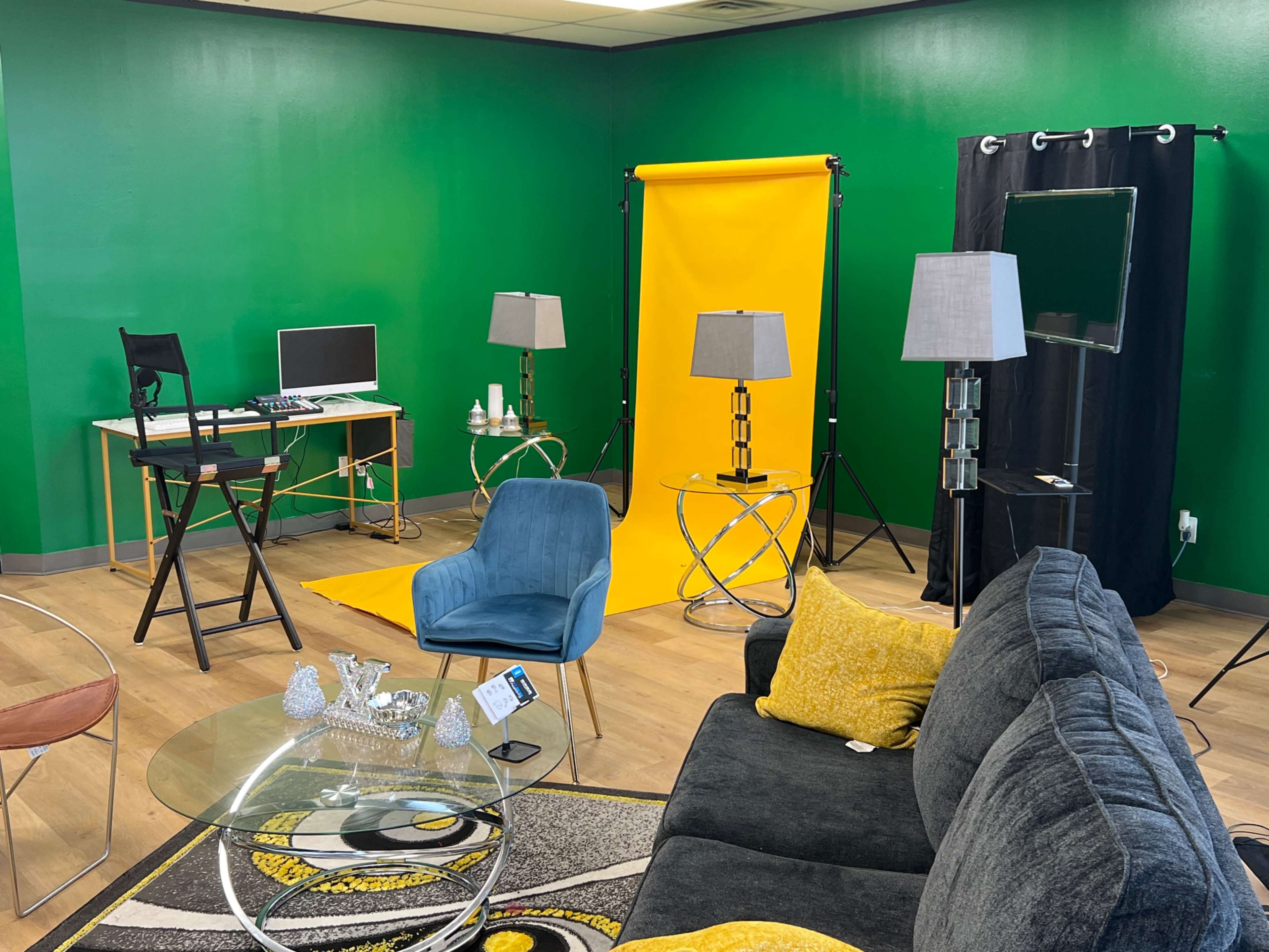 A studio setup featuring a green wall, yellow backdrop, a desktop computer, lighting equipment, a gray sofa, and a glass coffee table.