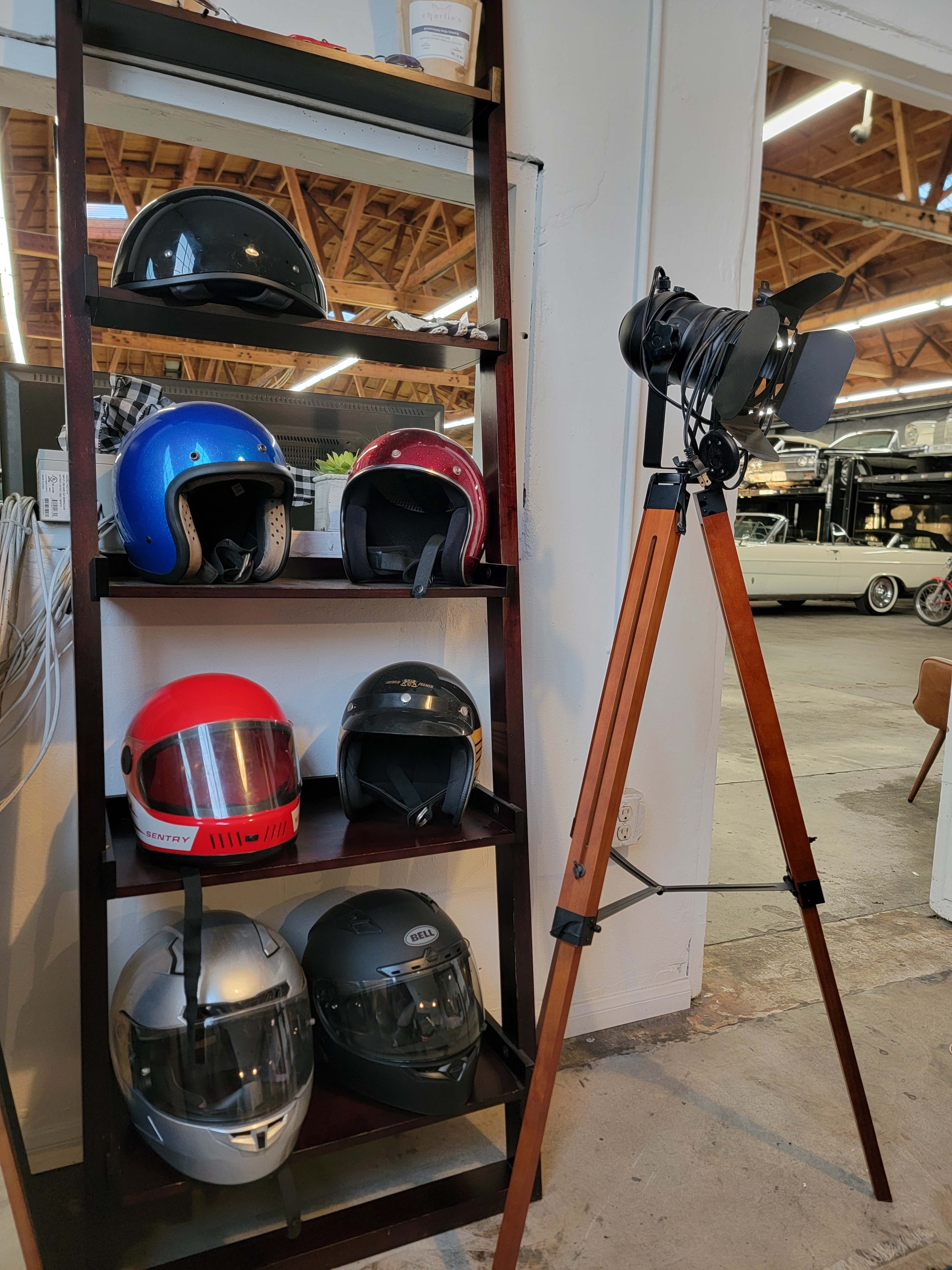 The image shows a wooden shelf displaying multiple motorcycle helmets of various colors, alongside a vintage camera on a tripod.