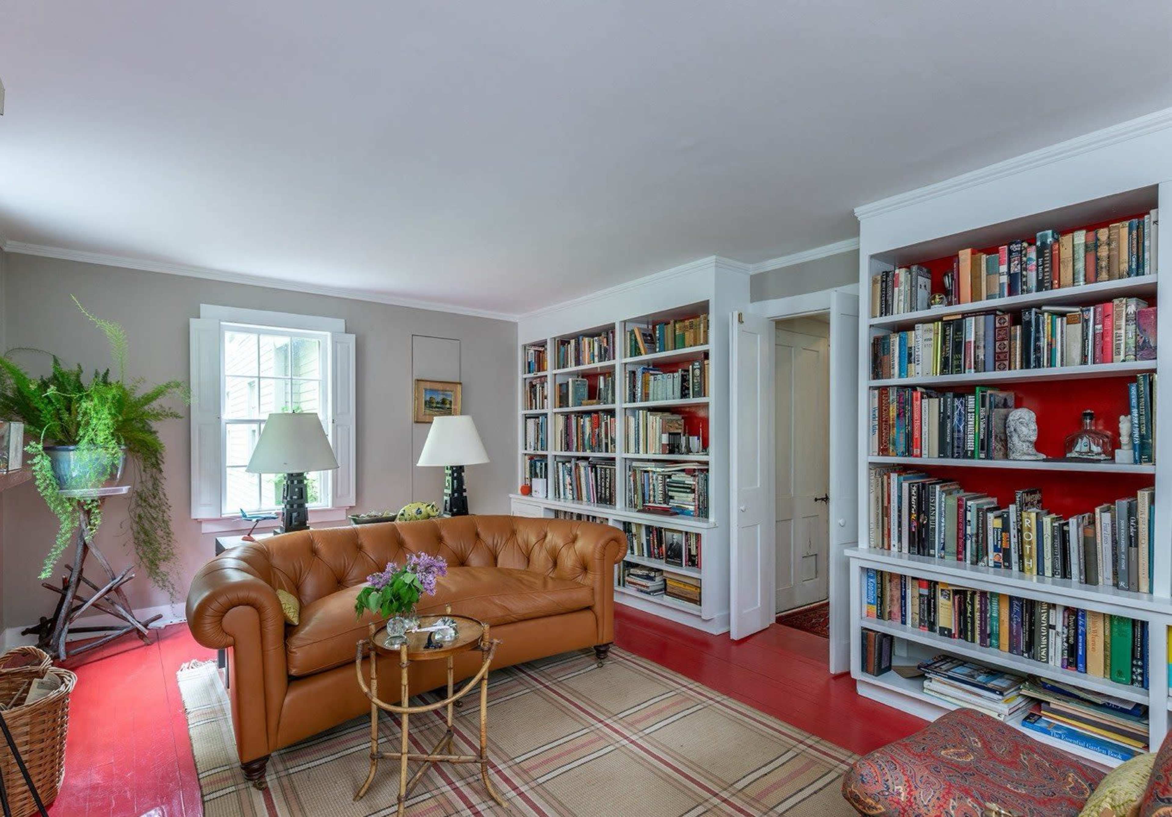The image shows a cozy living room featuring a brown leather couch, two lamps, bookshelves filled with books, and a small table with a vase of flowers.