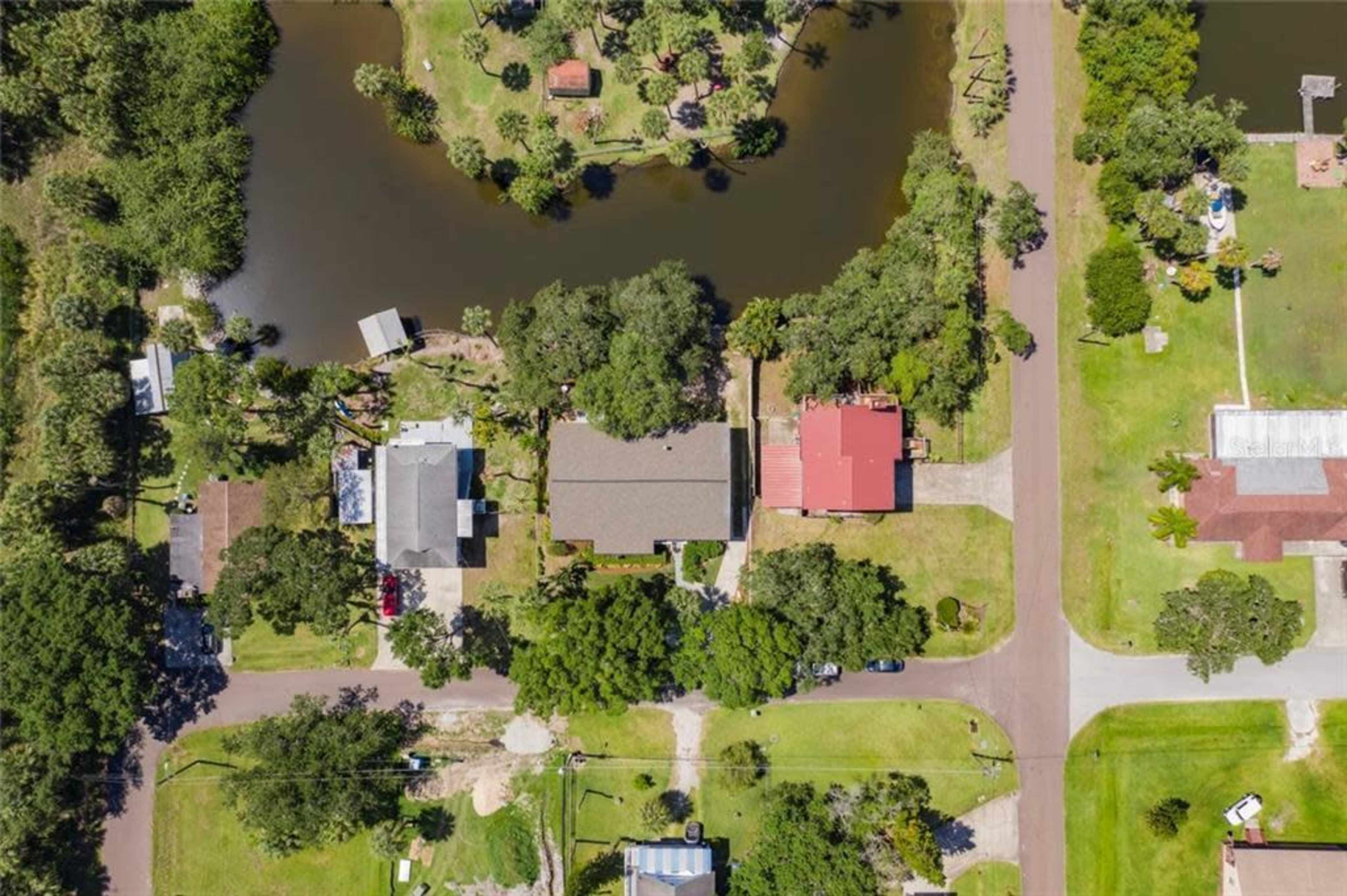 An aerial view shows a residential area featuring several houses, roads, and a small pond surrounded by greenery.