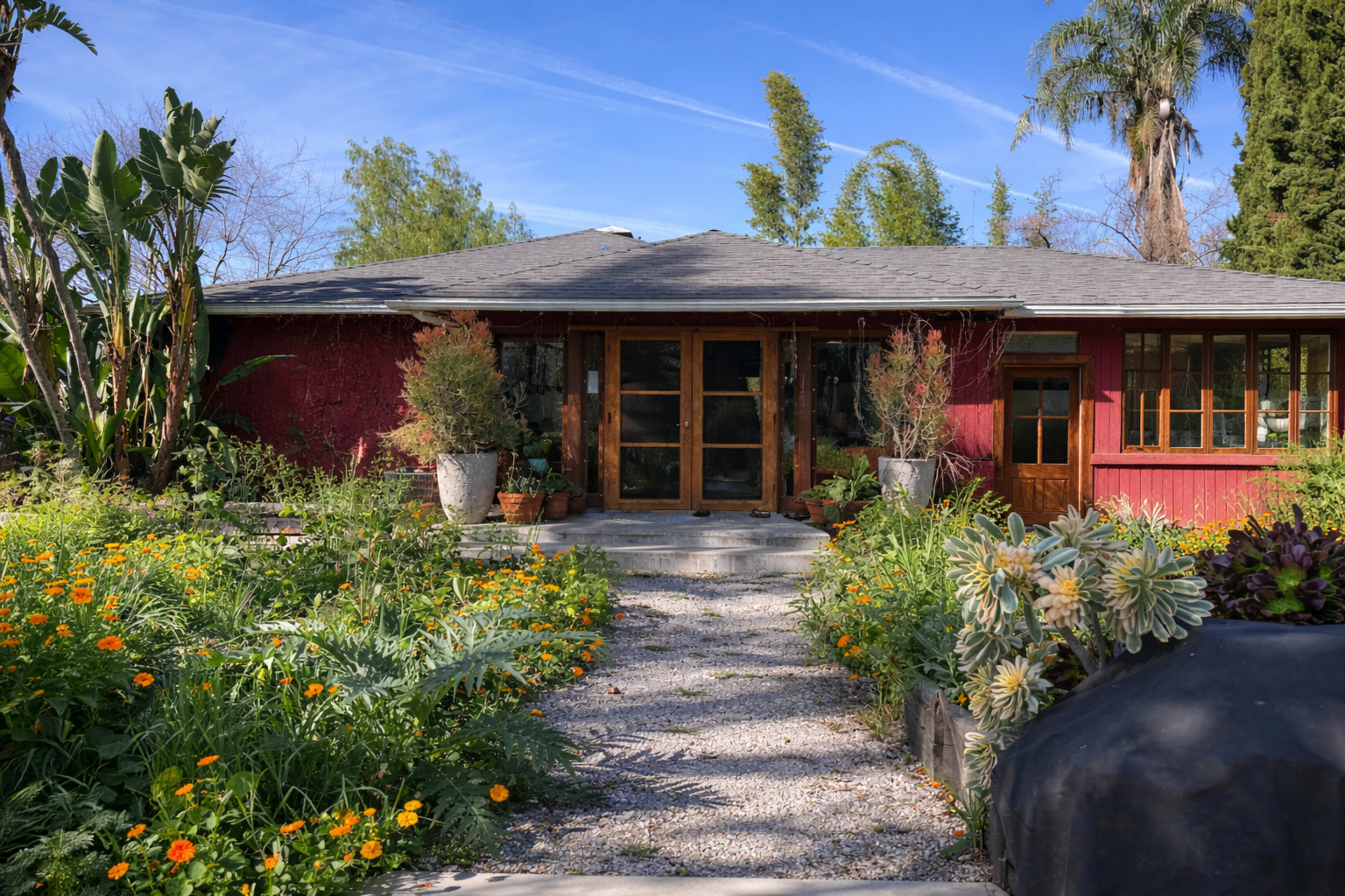The image shows a house with a red exterior, featuring a wide pathway lined with colorful flowers and plants, leading to a front door.