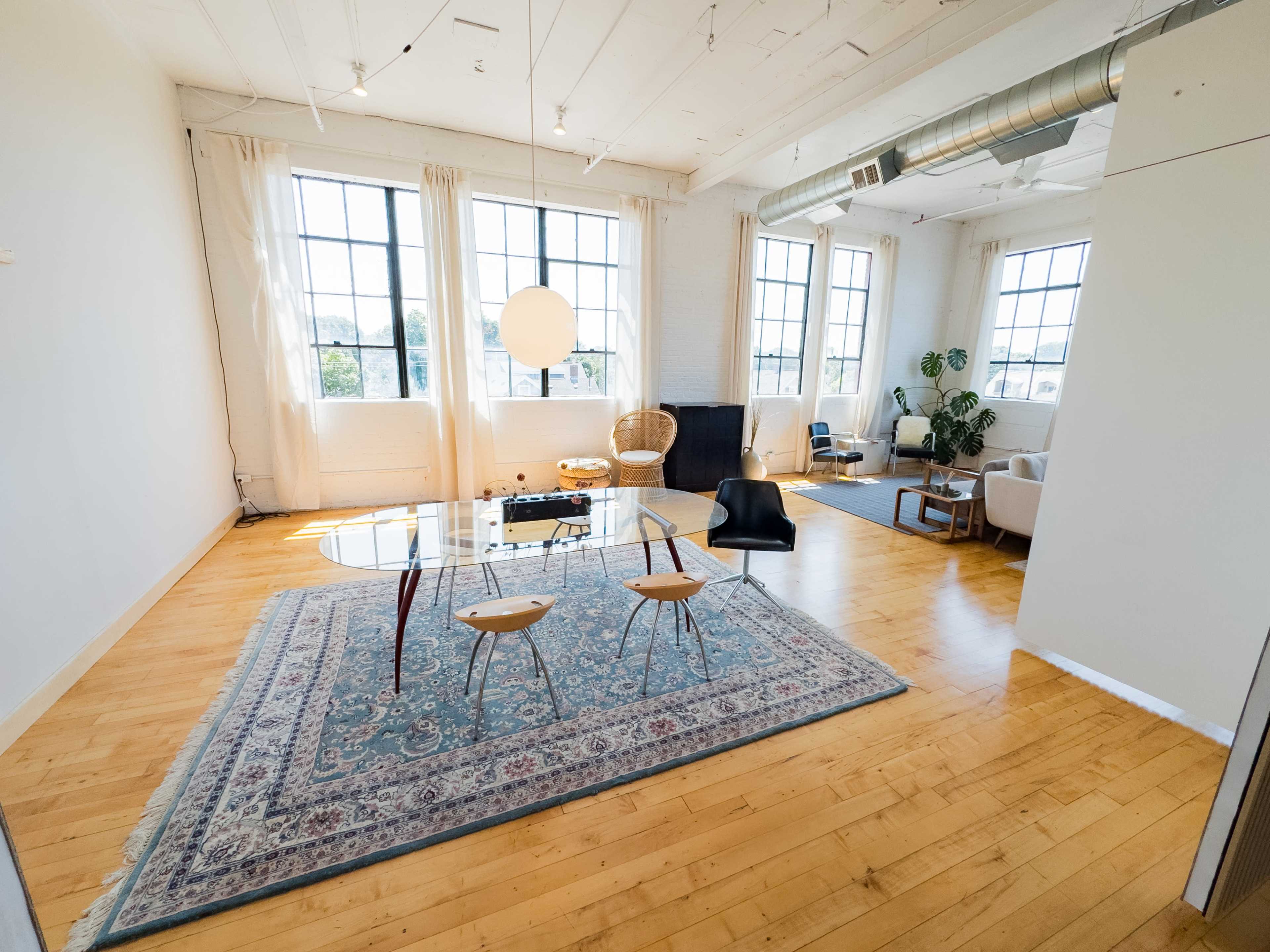 The image shows a spacious, brightly lit room featuring large windows, a glass dining table with two chairs, and various seating arrangements.