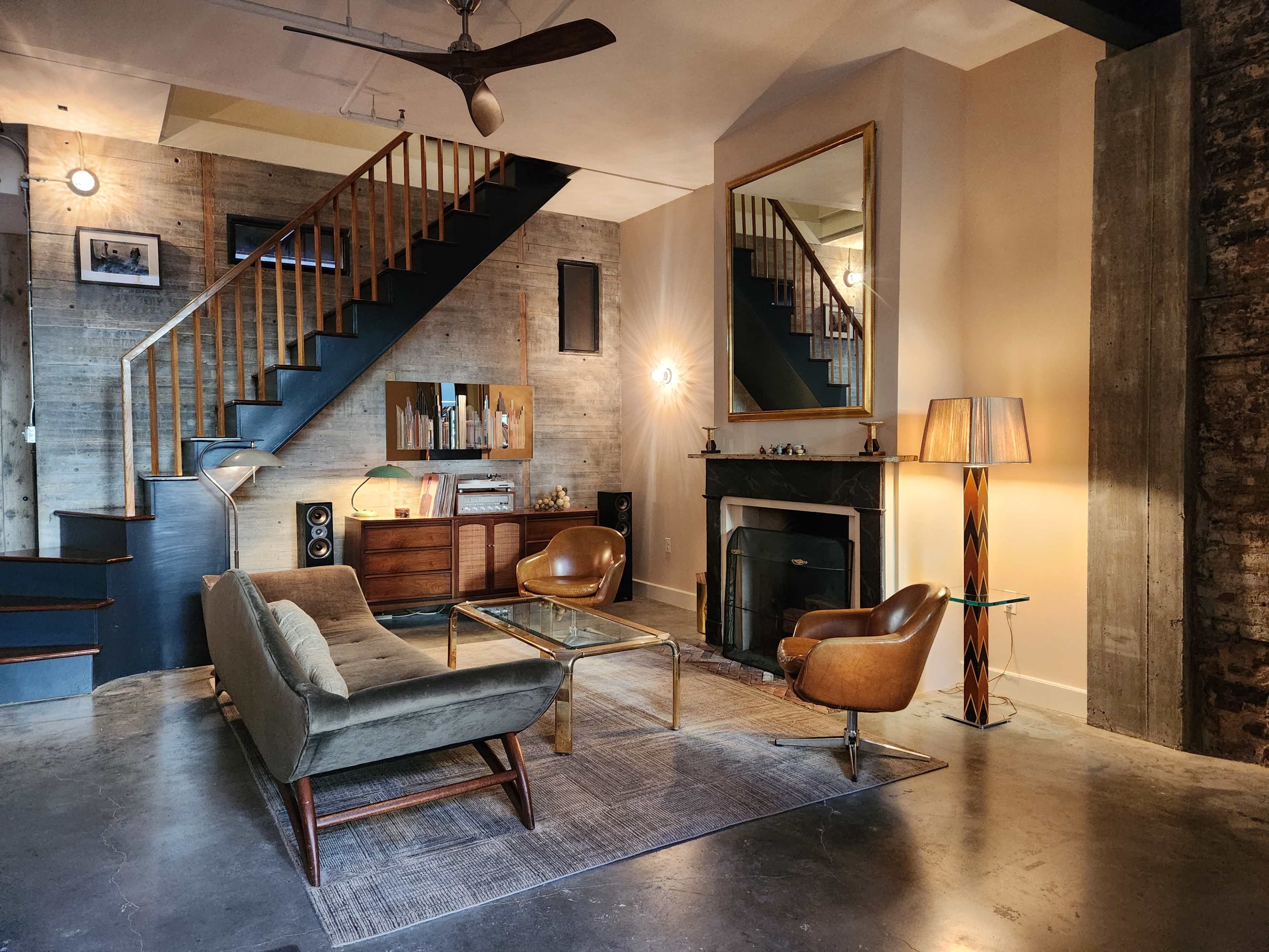 The image shows a stylish living room with a sofa, wooden furniture, a fireplace, and a staircase in the background.