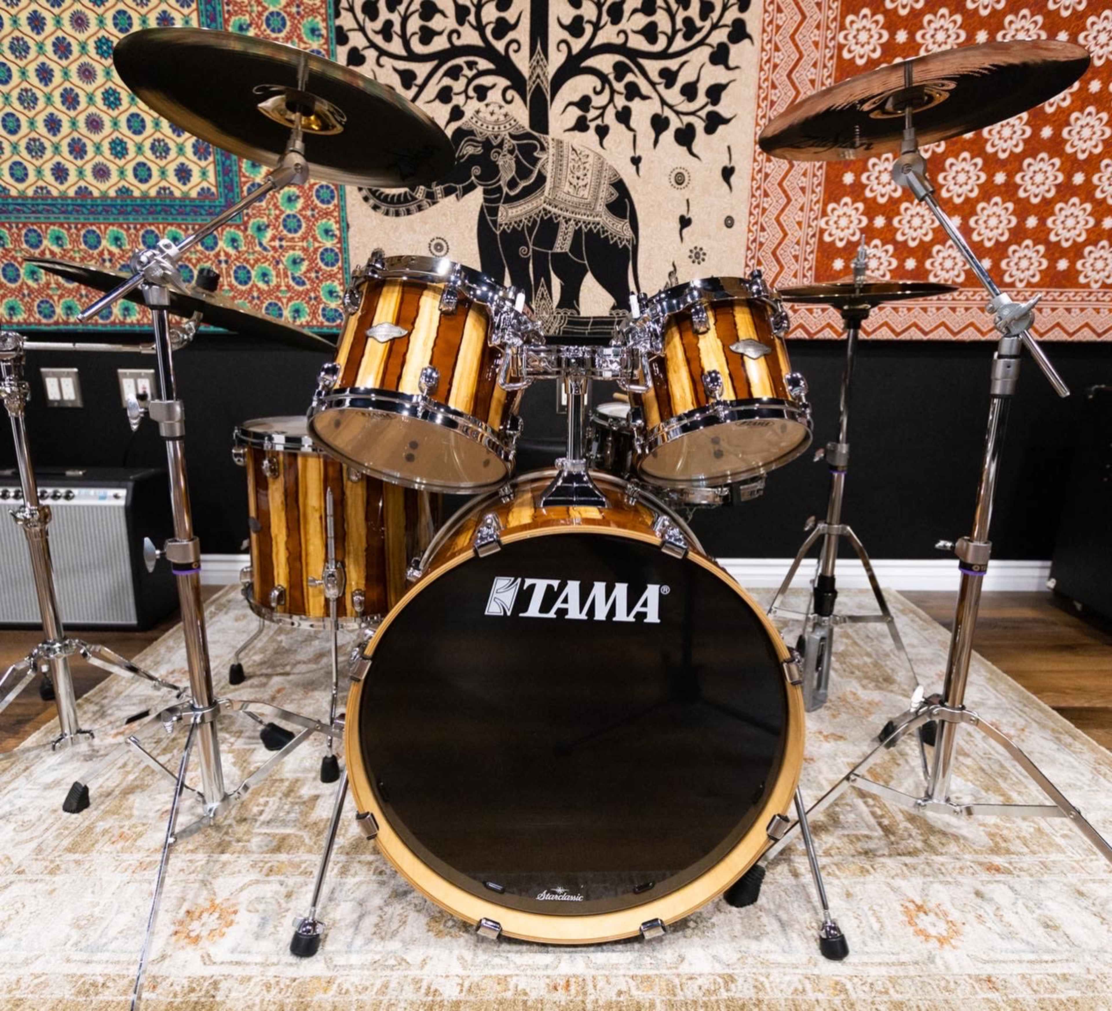 The image shows a Tama drum set with a natural wood finish, positioned on a decorative rug and surrounded by colorful wall tapestries.