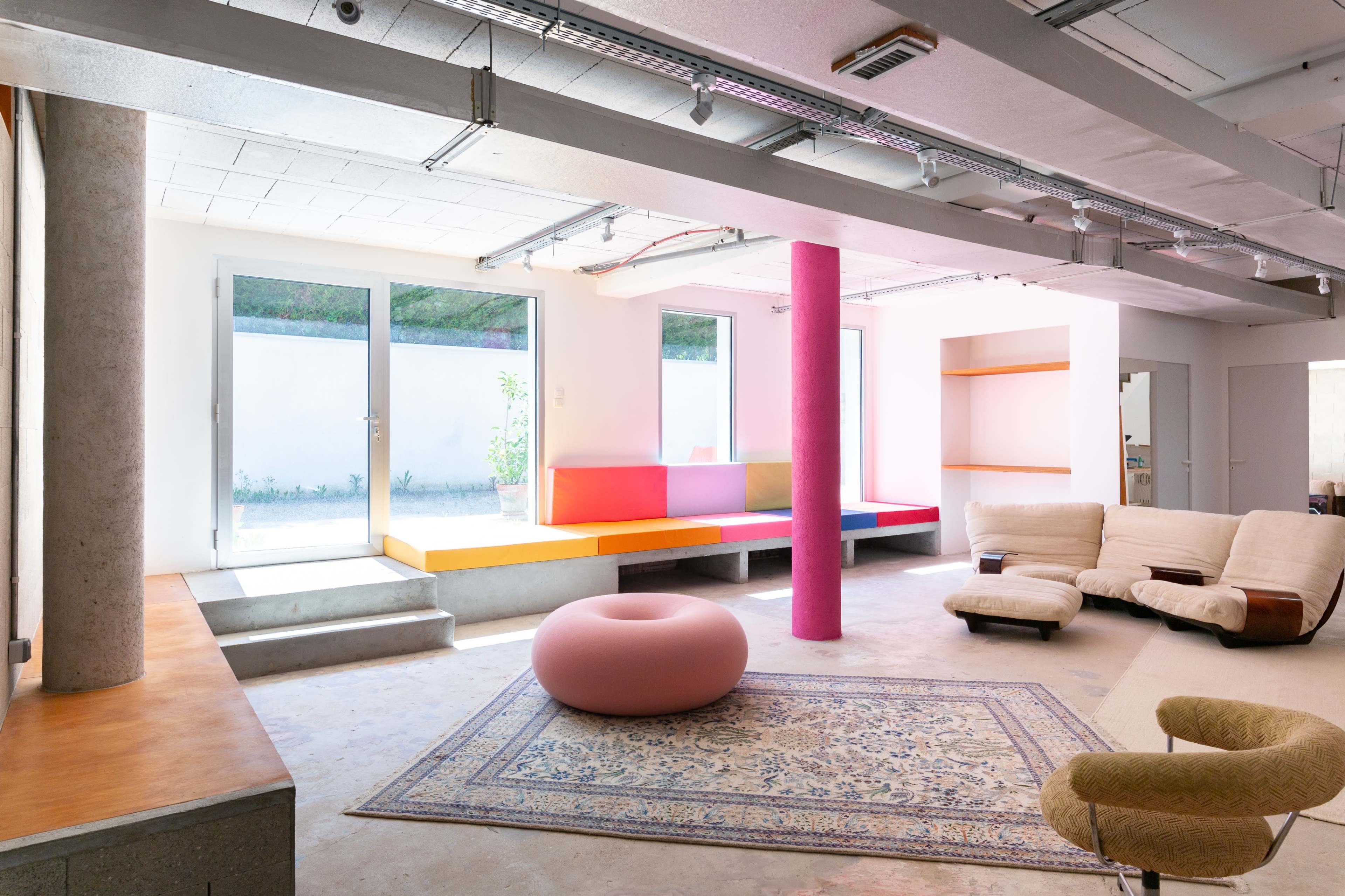 The image shows a spacious, modern interior featuring a colorful seating area, large windows, and a patterned rug.