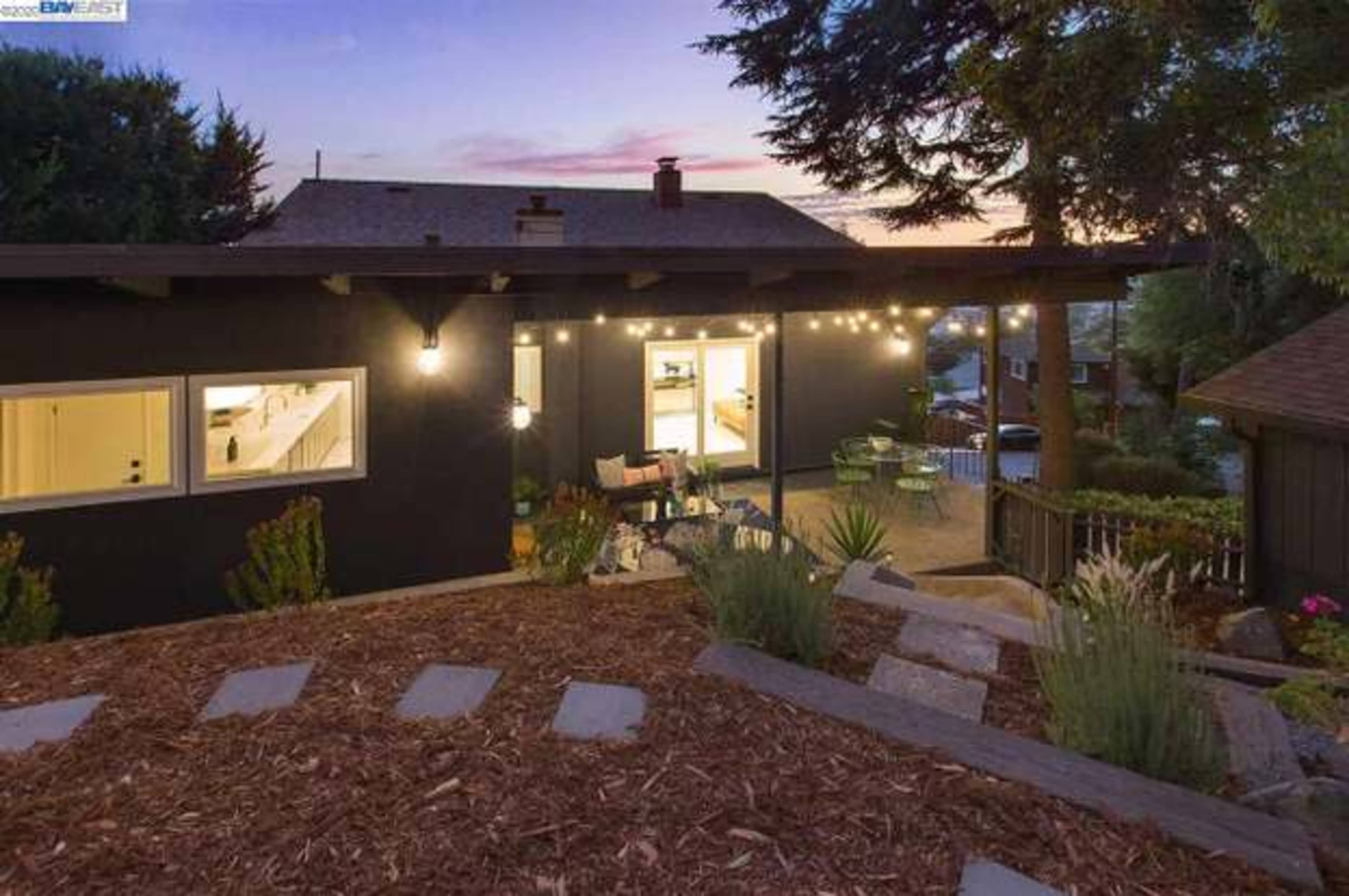 A hillside residential backyard with a modern house, string lights, and a patio area overlooking the landscape at sunset.
