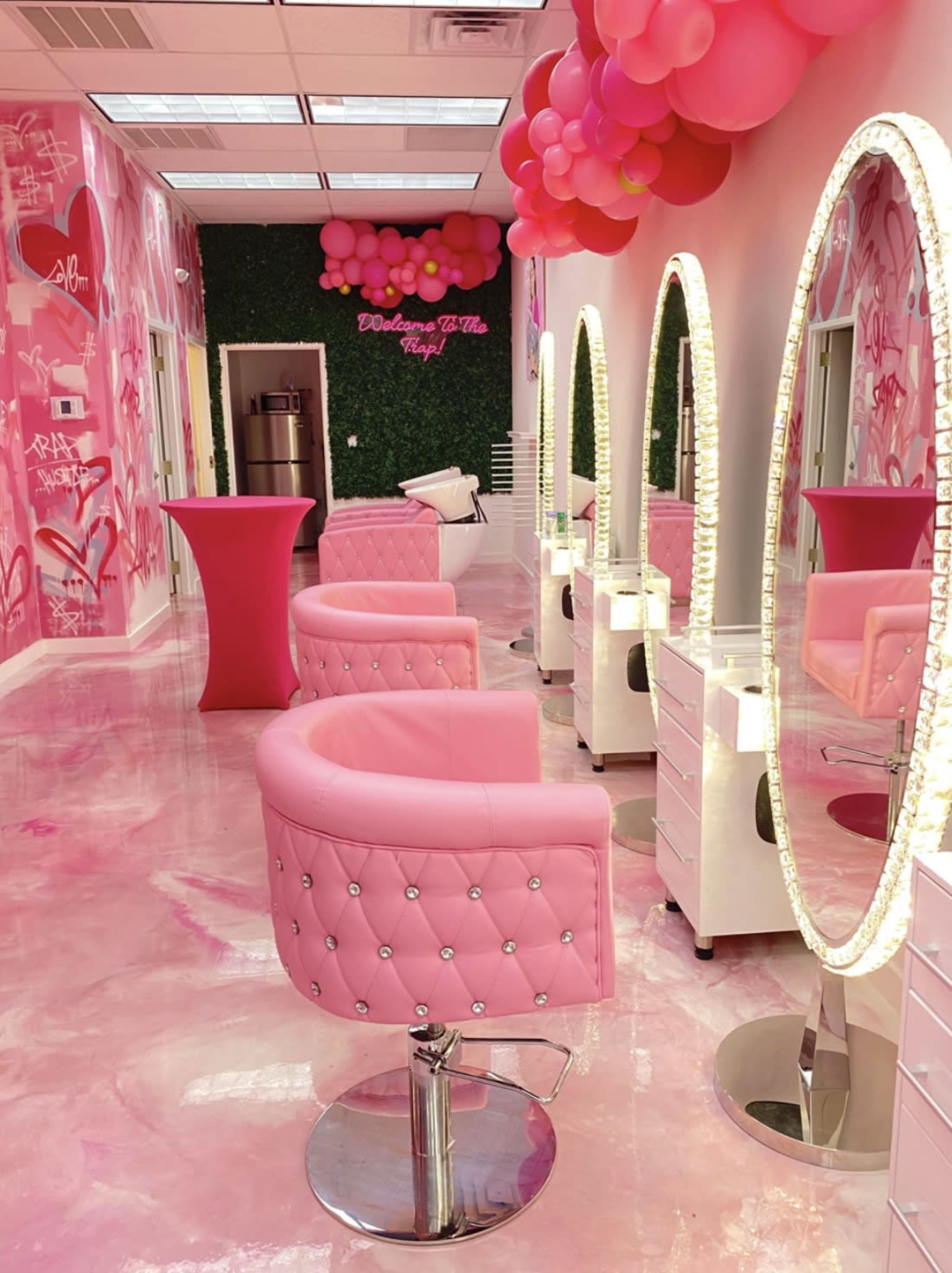 A brightly lit salon with pink furniture, decorative wall art, and modern styling stations arranged along a glossy floor.