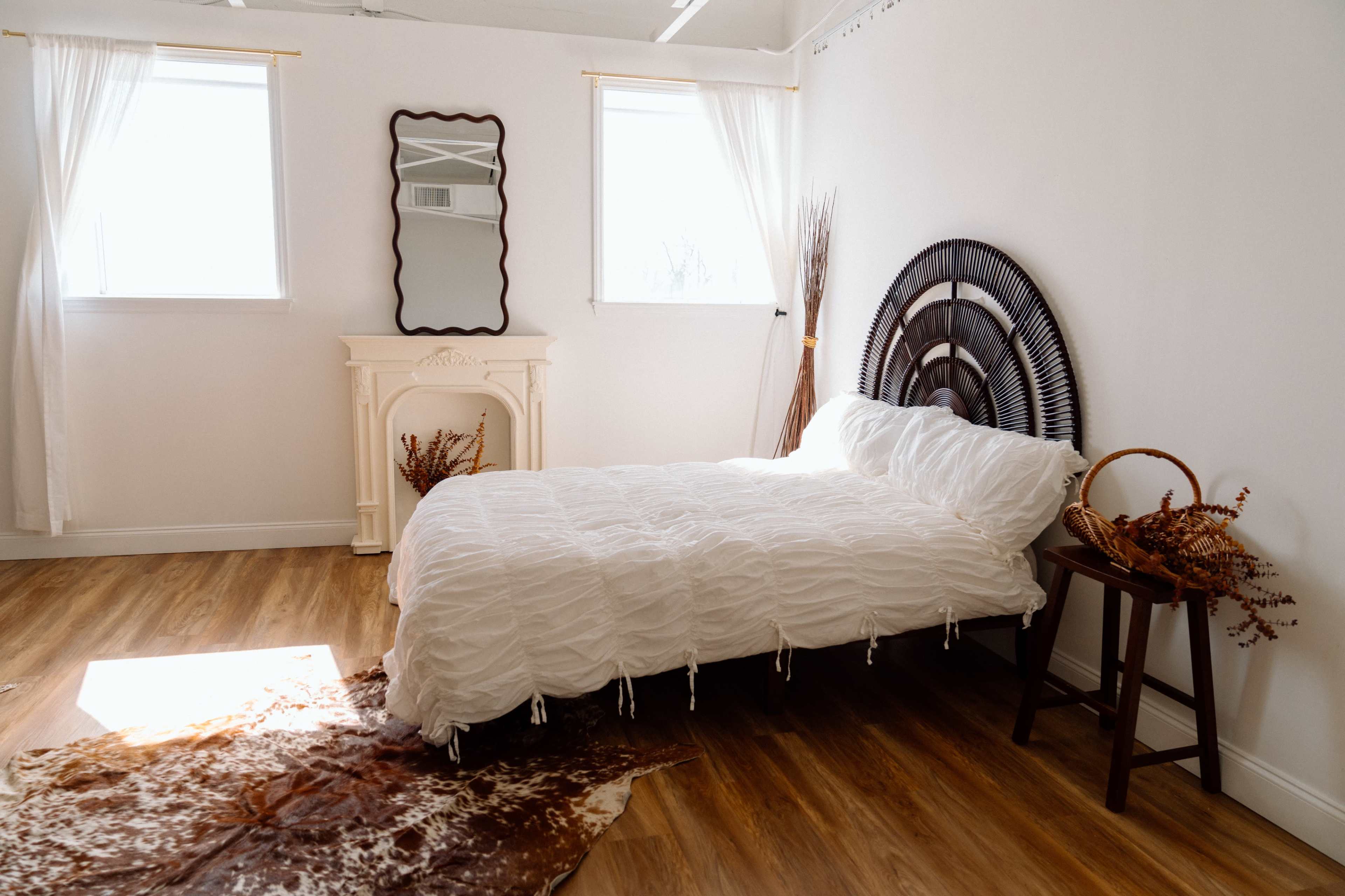 A minimalist bedroom features a bed with a textured white coverlet, a decorative mirror on the wall, and wooden flooring, complemented by natural light from two windows.
