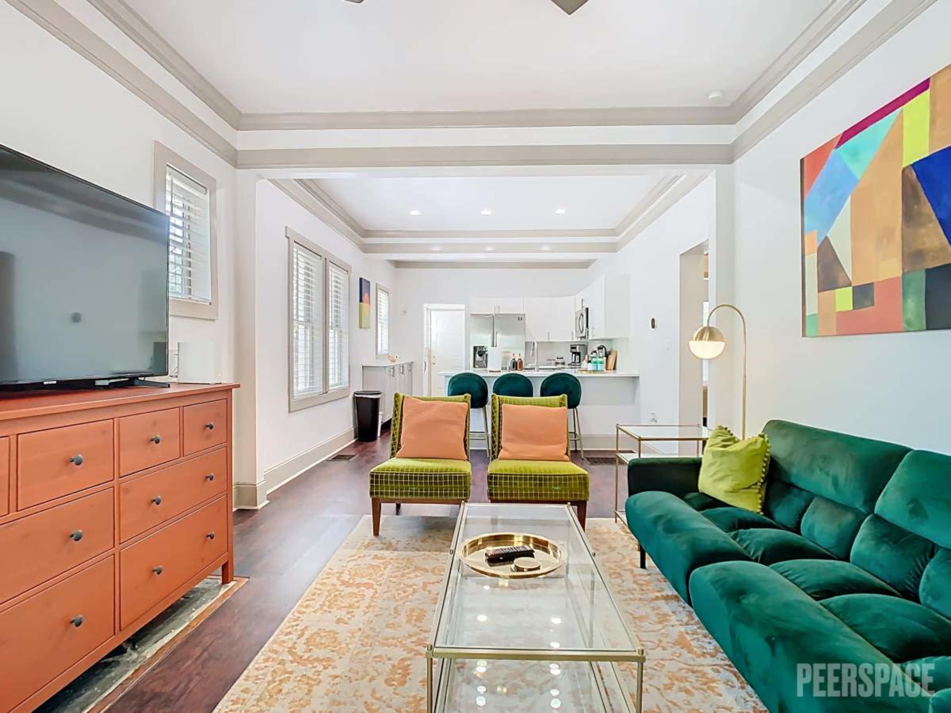 The image shows a living room with a green velvet sofa, orange armchairs, a television, and a kitchen area visible in the background.