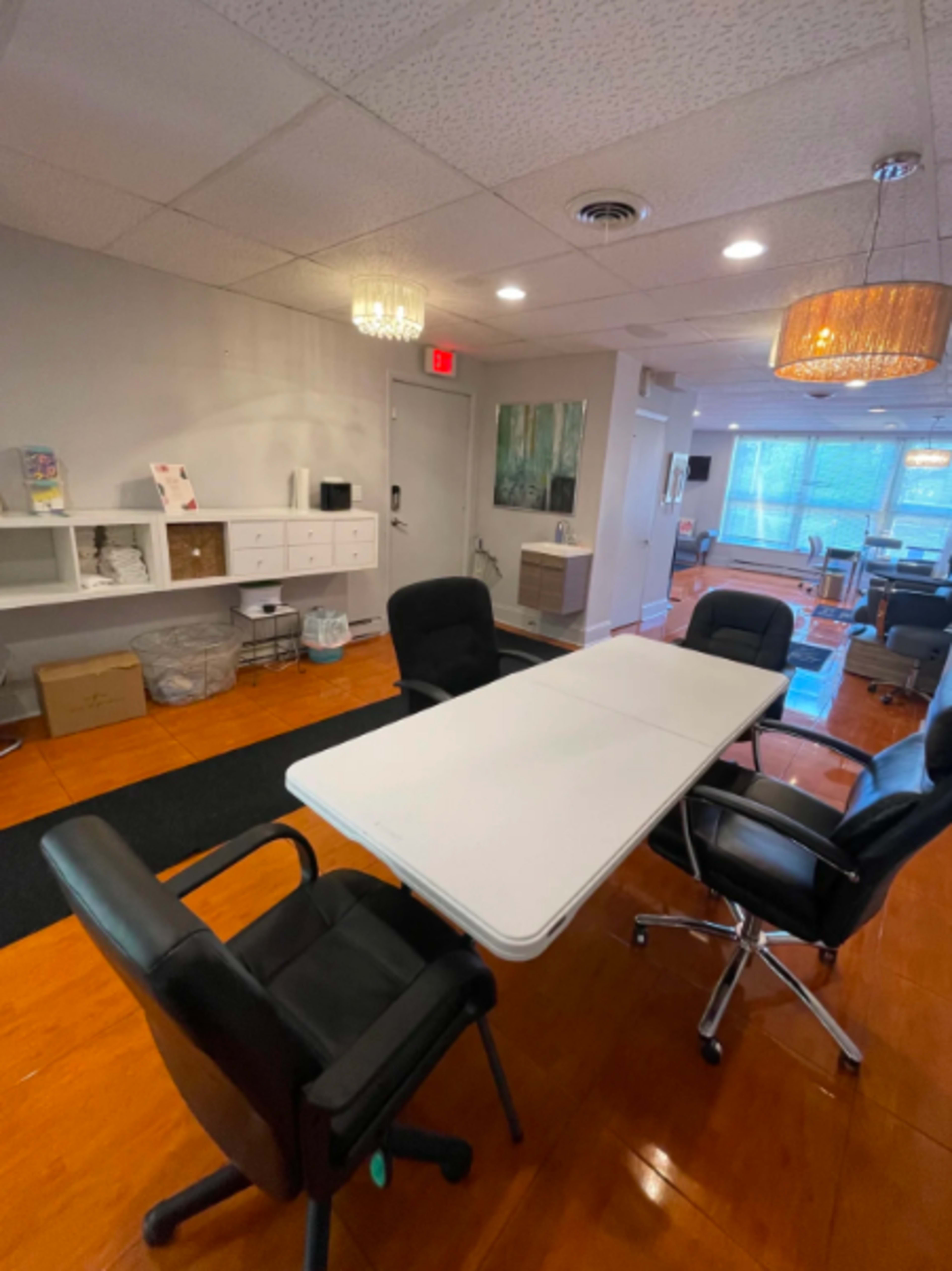 The image shows a modern office space with a central conference table surrounded by four black chairs, featuring wooden flooring and various office furnishings along the walls.