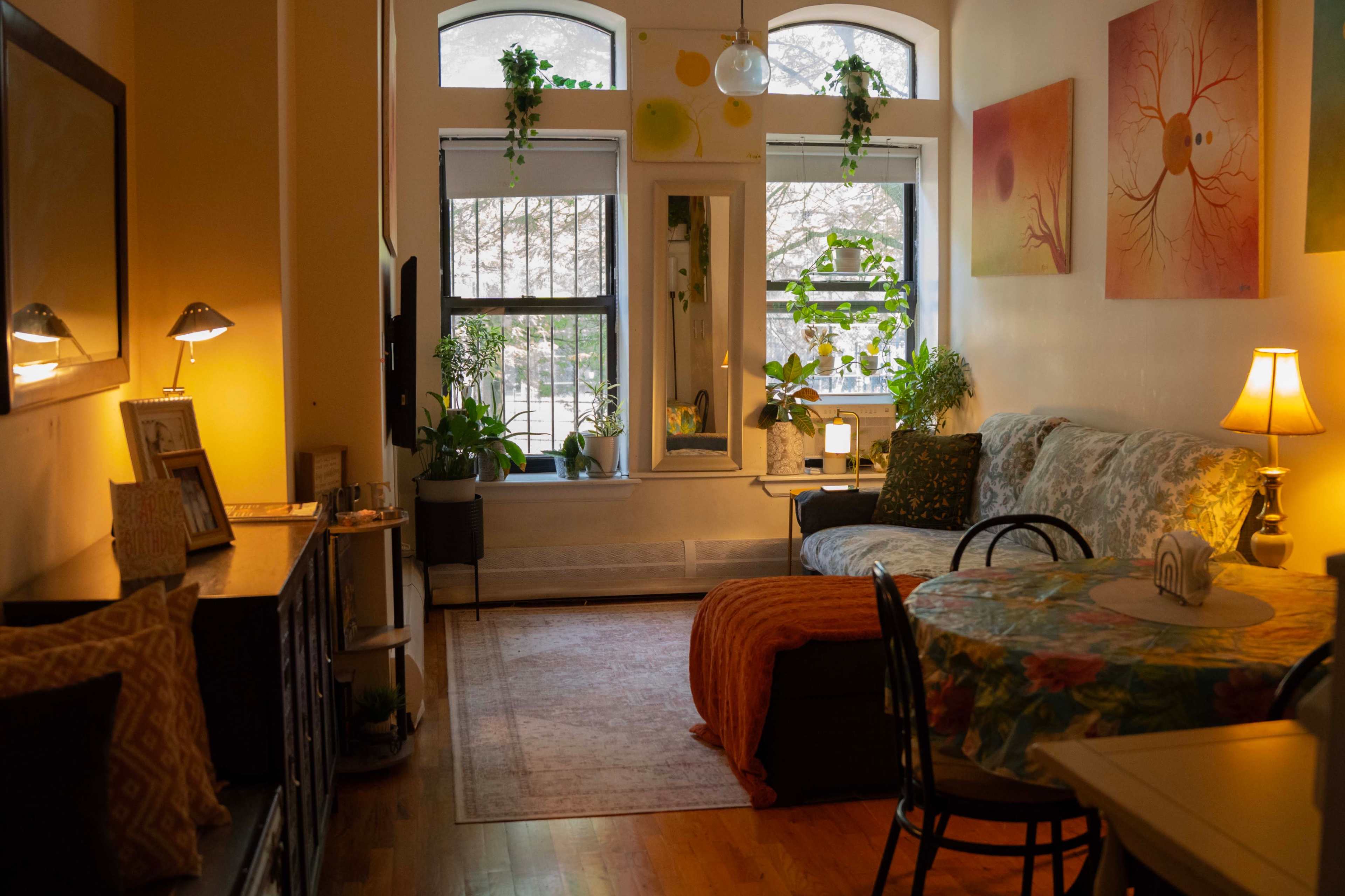 The image shows a cozy living space with a sofa, a table, and several plants near large windows, all illuminated by warm lighting.