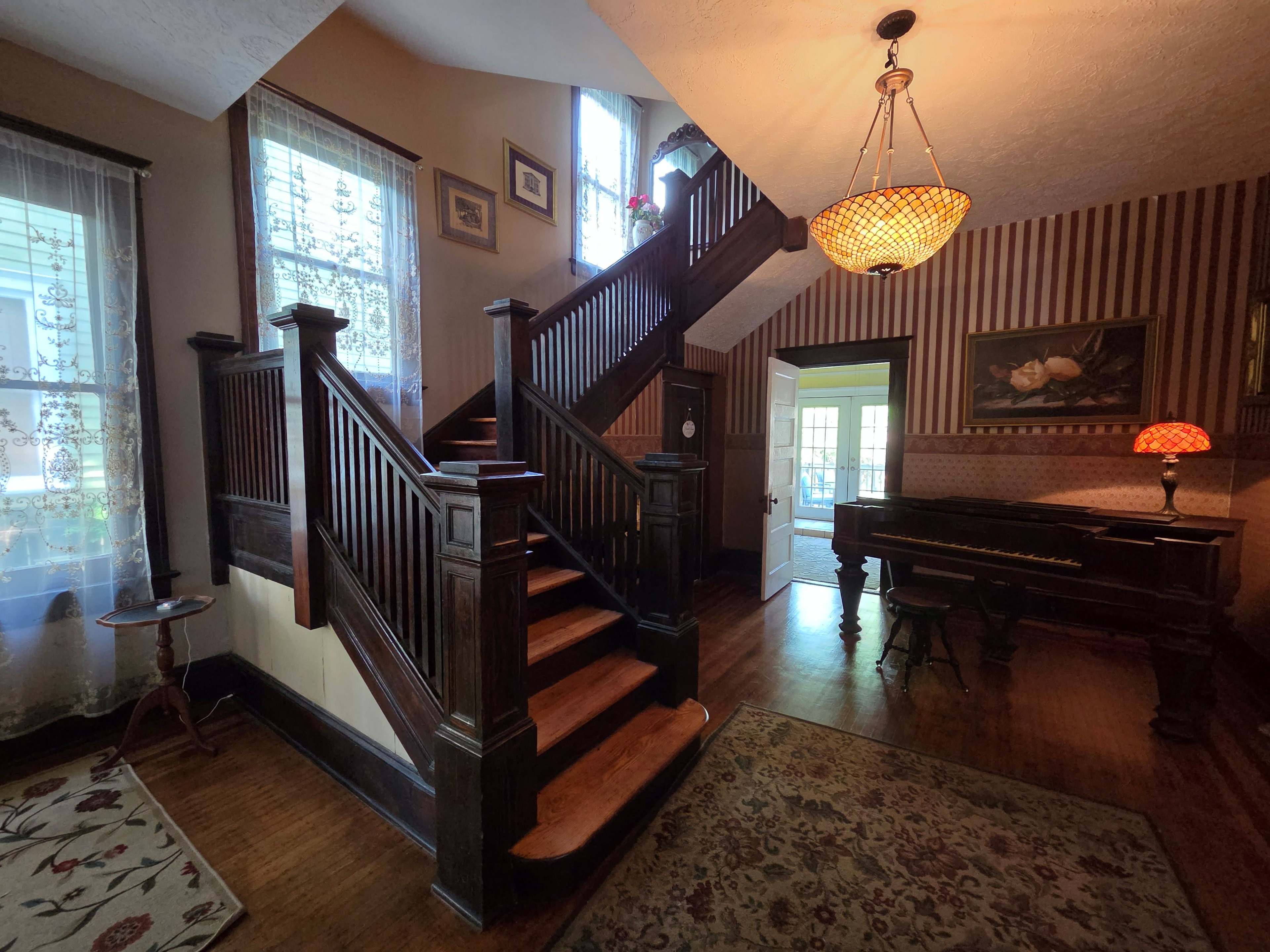 The image shows an interior foyer with a wooden staircase, a vintage piano, and patterned wallpaper.