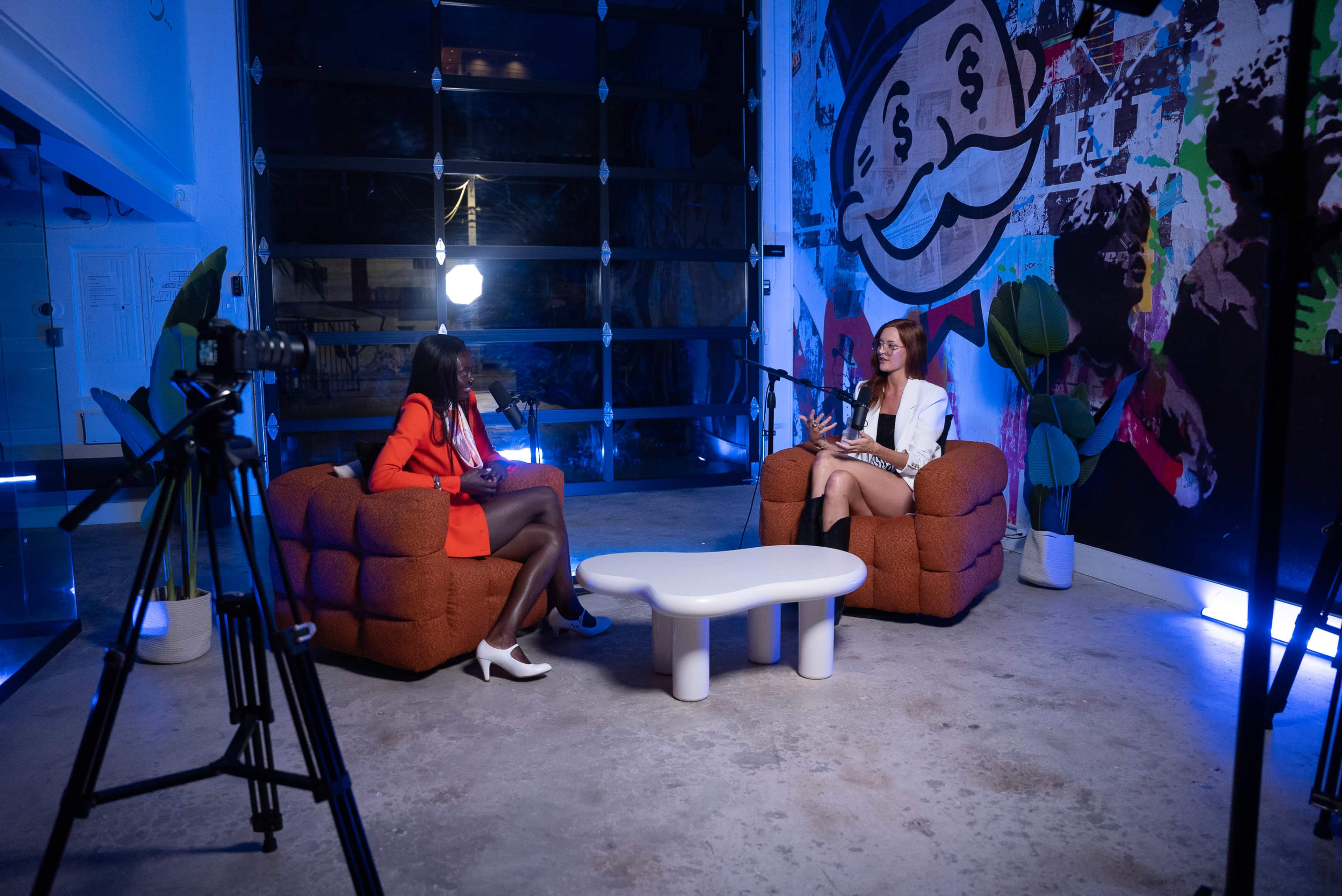 Two women are seated in an interview setup on brown upholstered chairs with a round white table between them, surrounded by a colorful mural and camera equipment in a well-lit space.