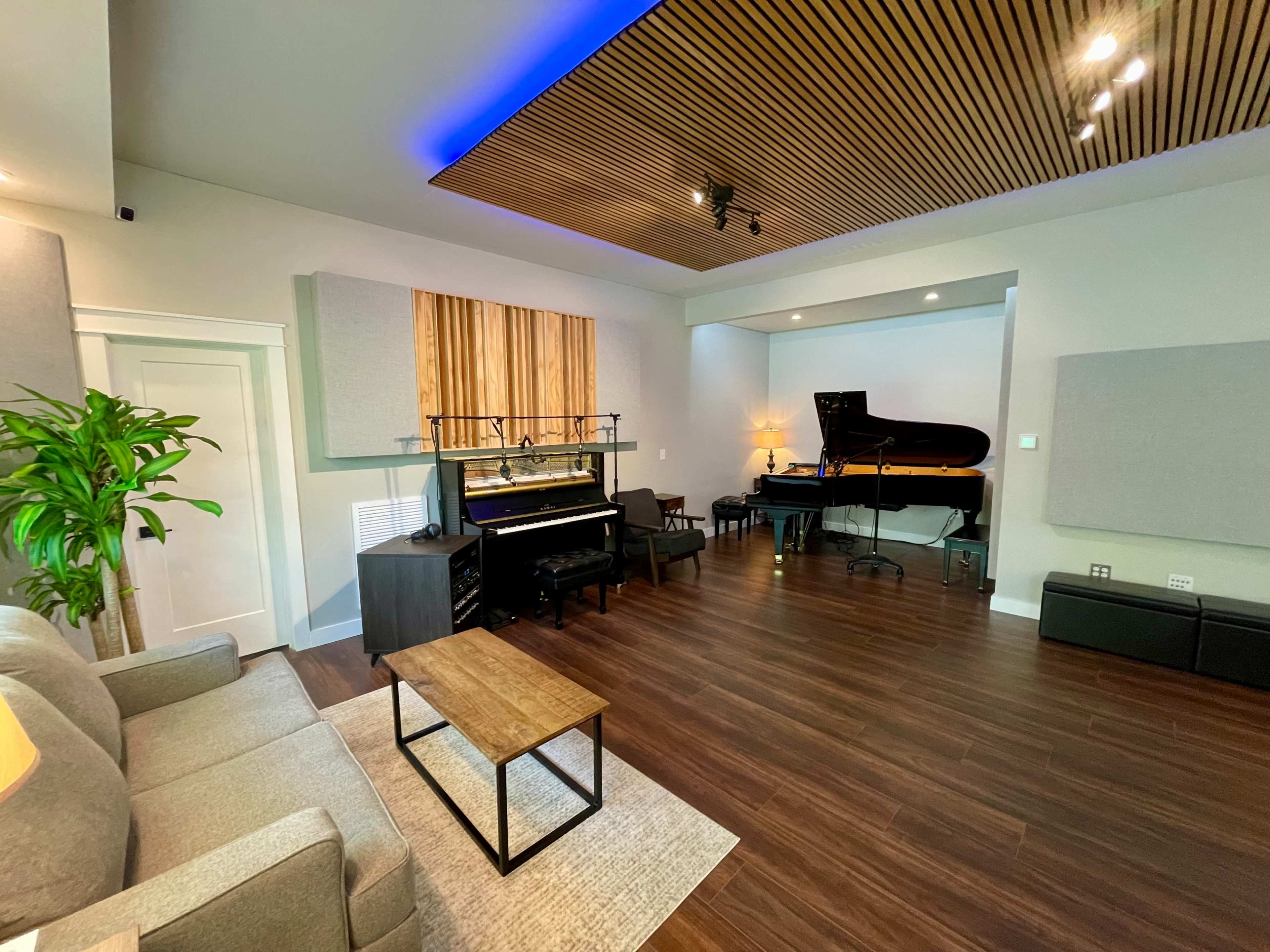 A music studio featuring a grand piano, a black upright piano, a sofa, and wooden acoustic panels on the ceiling.