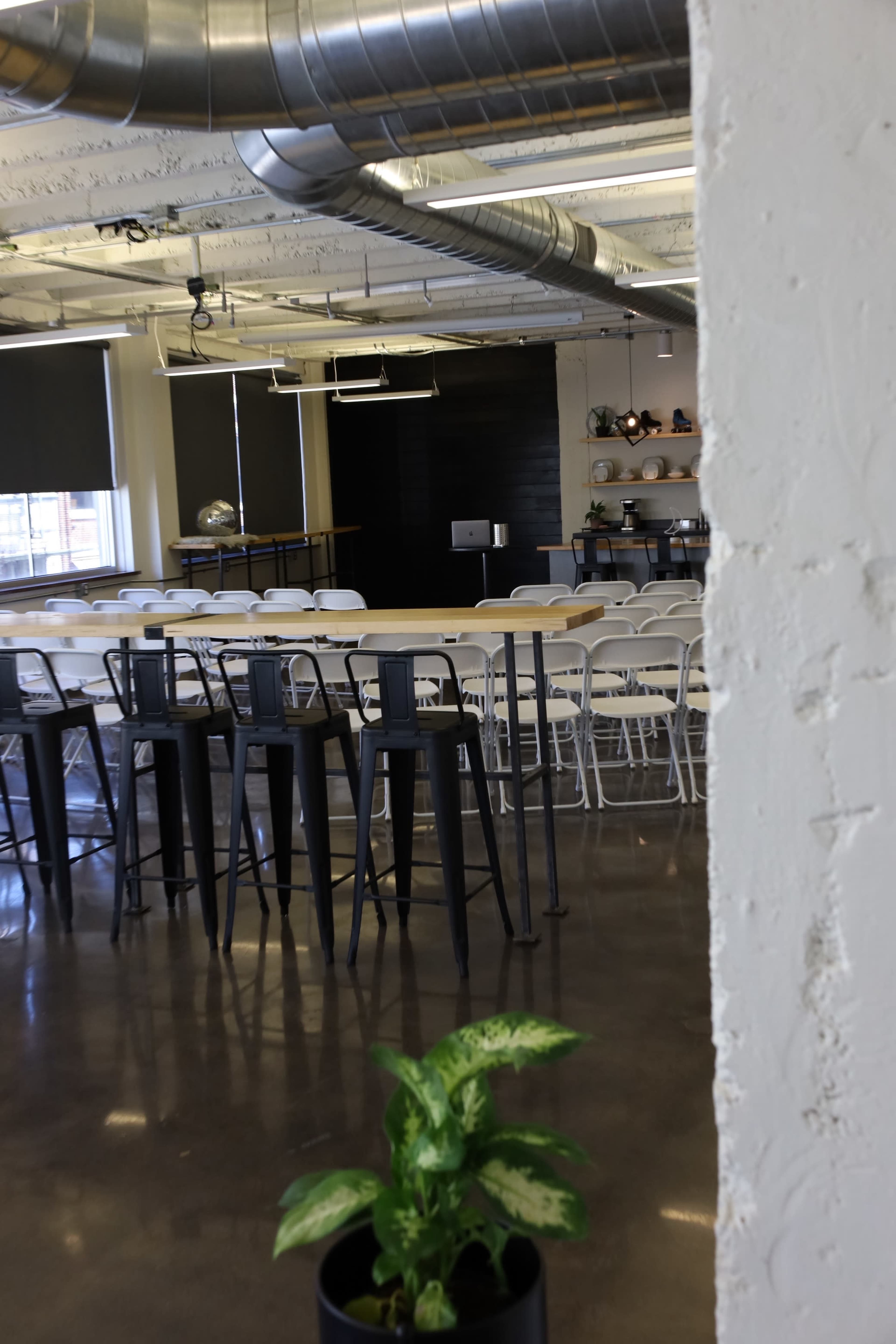 A modern, industrial-style meeting room with rows of white chairs, high-top tables, and large windows.