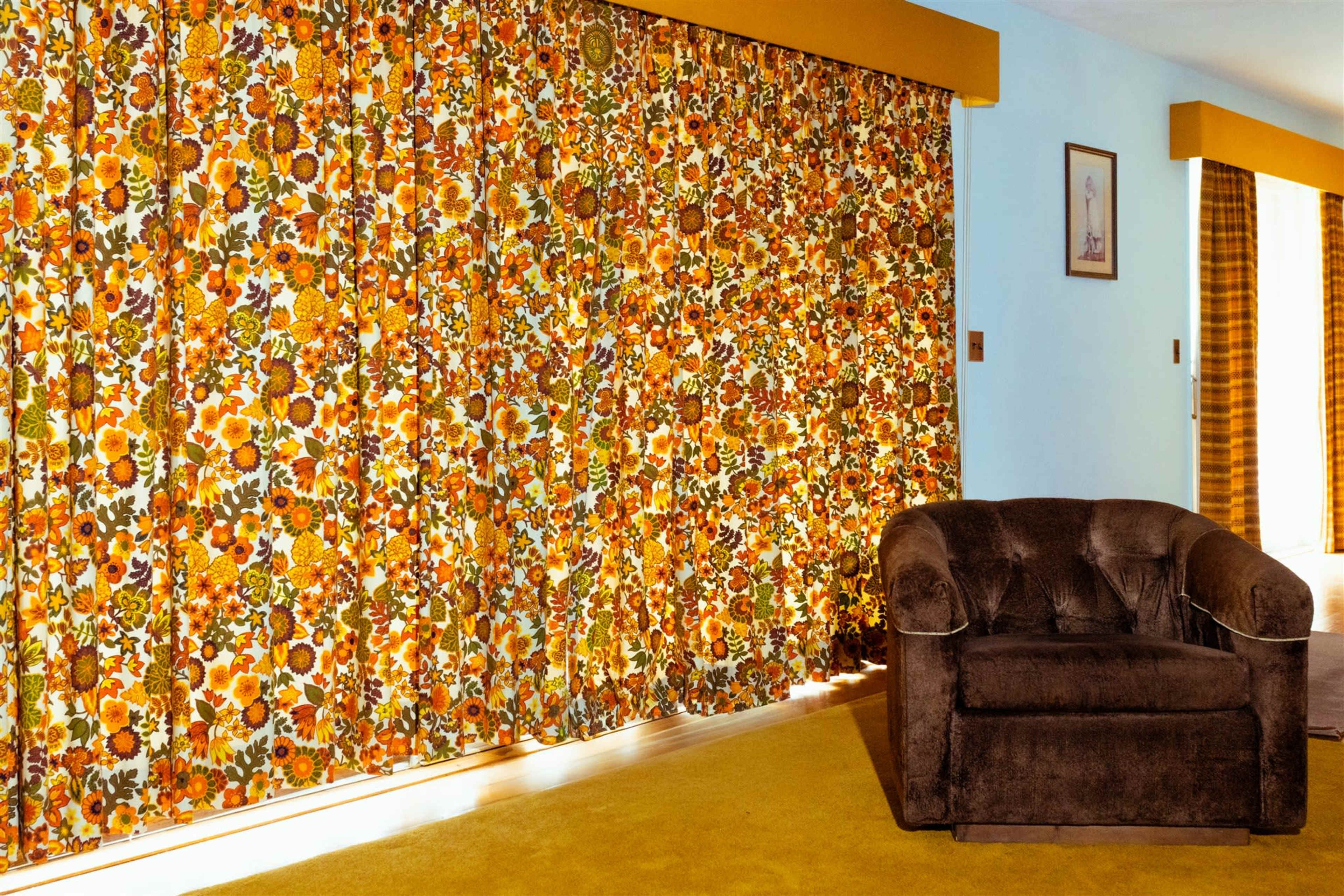 The image shows a room with vibrant, floral-patterned curtains, a brown armchair, and yellow carpeting.