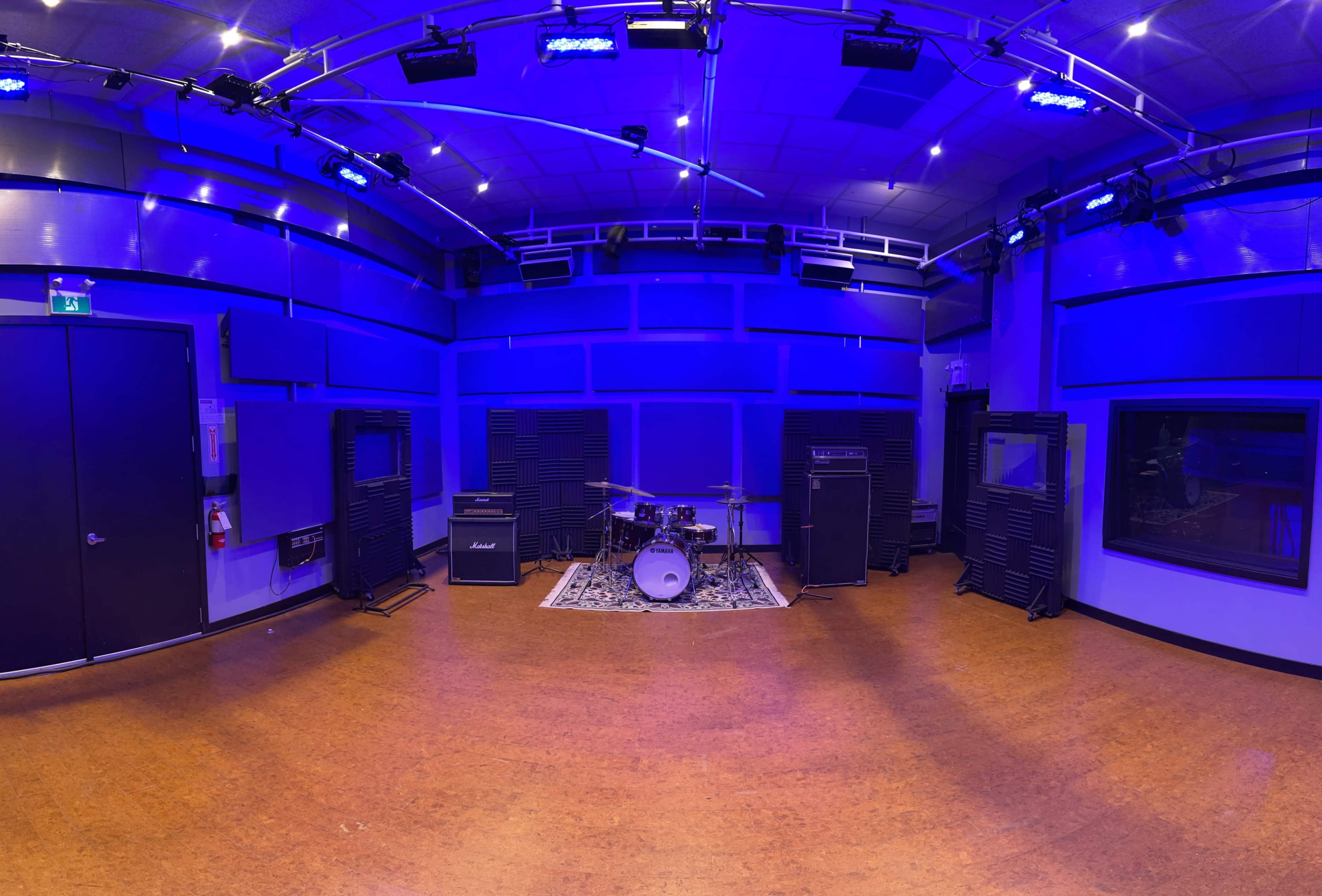 The image shows a music studio with a drum set and amplifiers arranged against blue-lit walls.