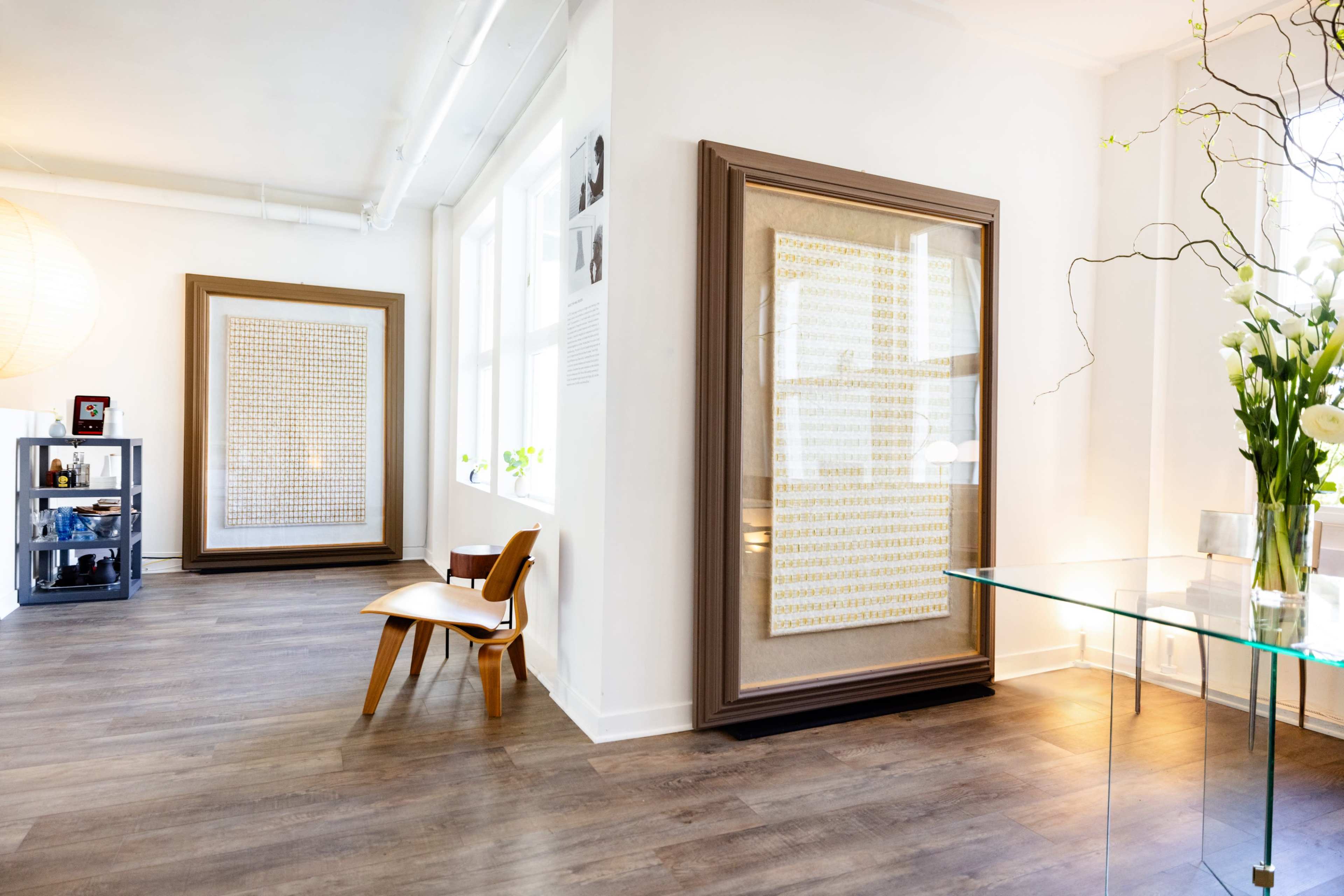 The image shows a bright, modern gallery space with two large framed artworks on the walls, a clear glass table, and a wooden chair.