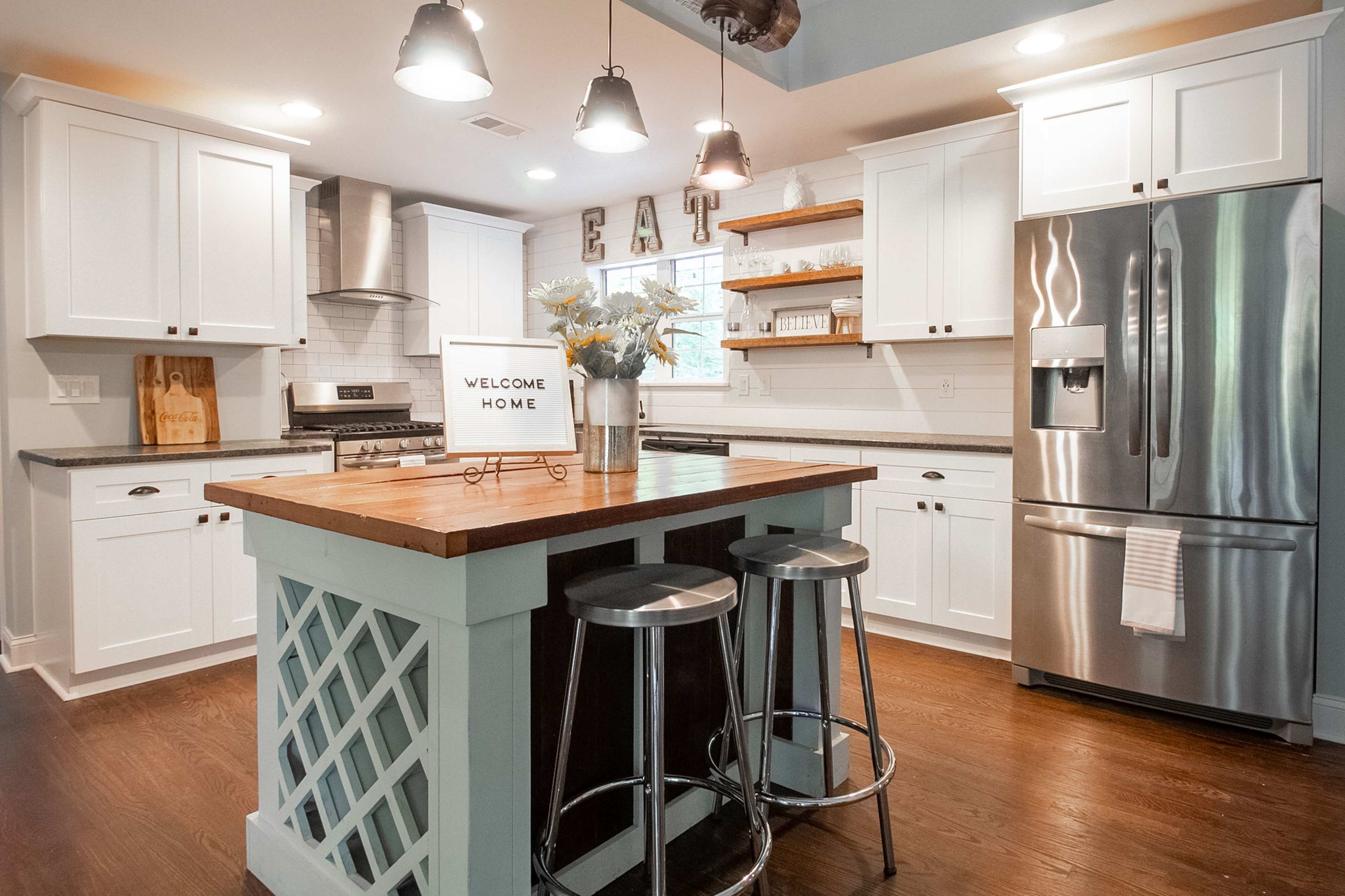 The image shows a modern kitchen with white cabinetry, a wooden island, stainless steel appliances, and bar stools.