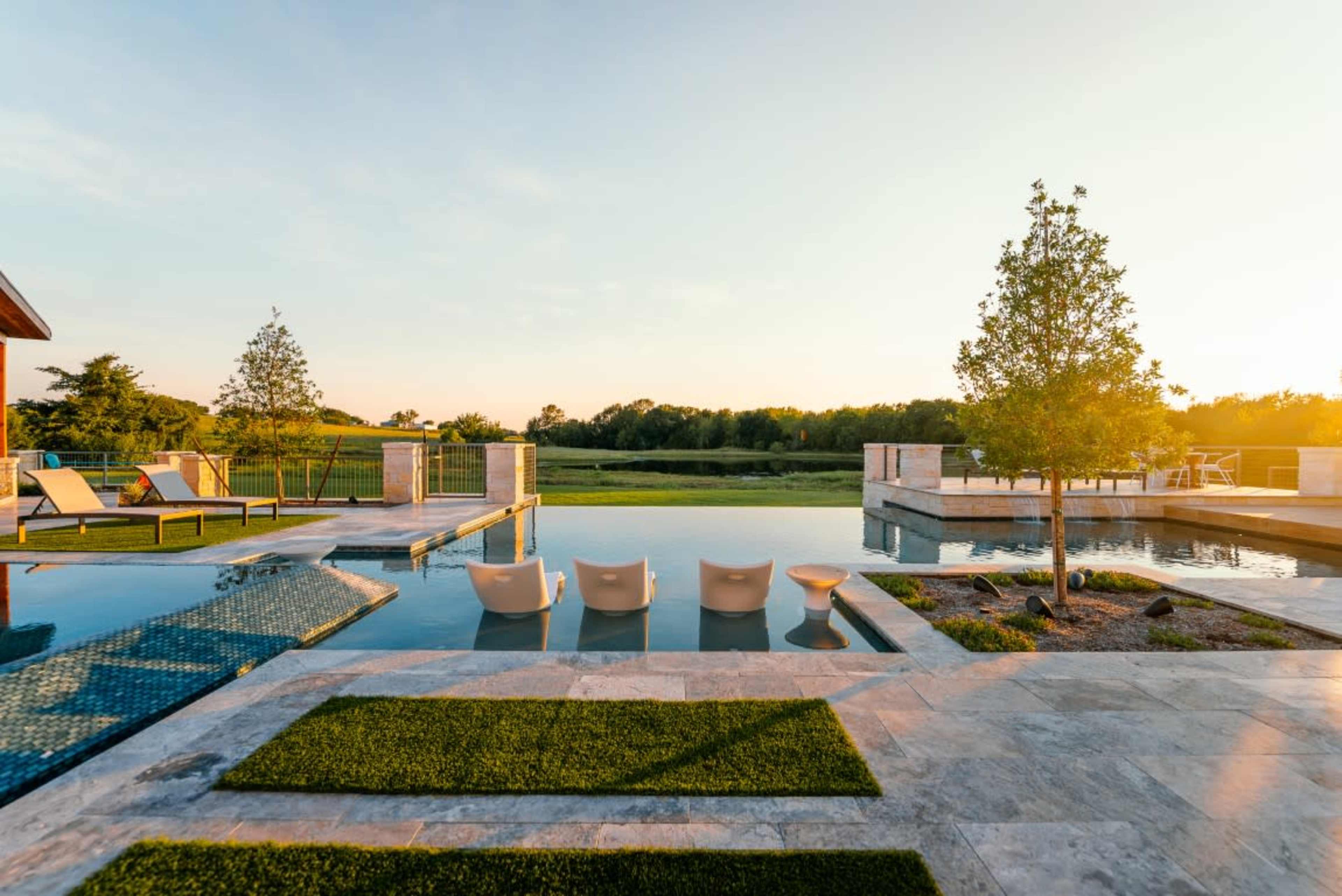 The image shows a modern outdoor space featuring a swimming pool with lounge chairs, surrounded by grass and trees, under a clear sky at sunset.