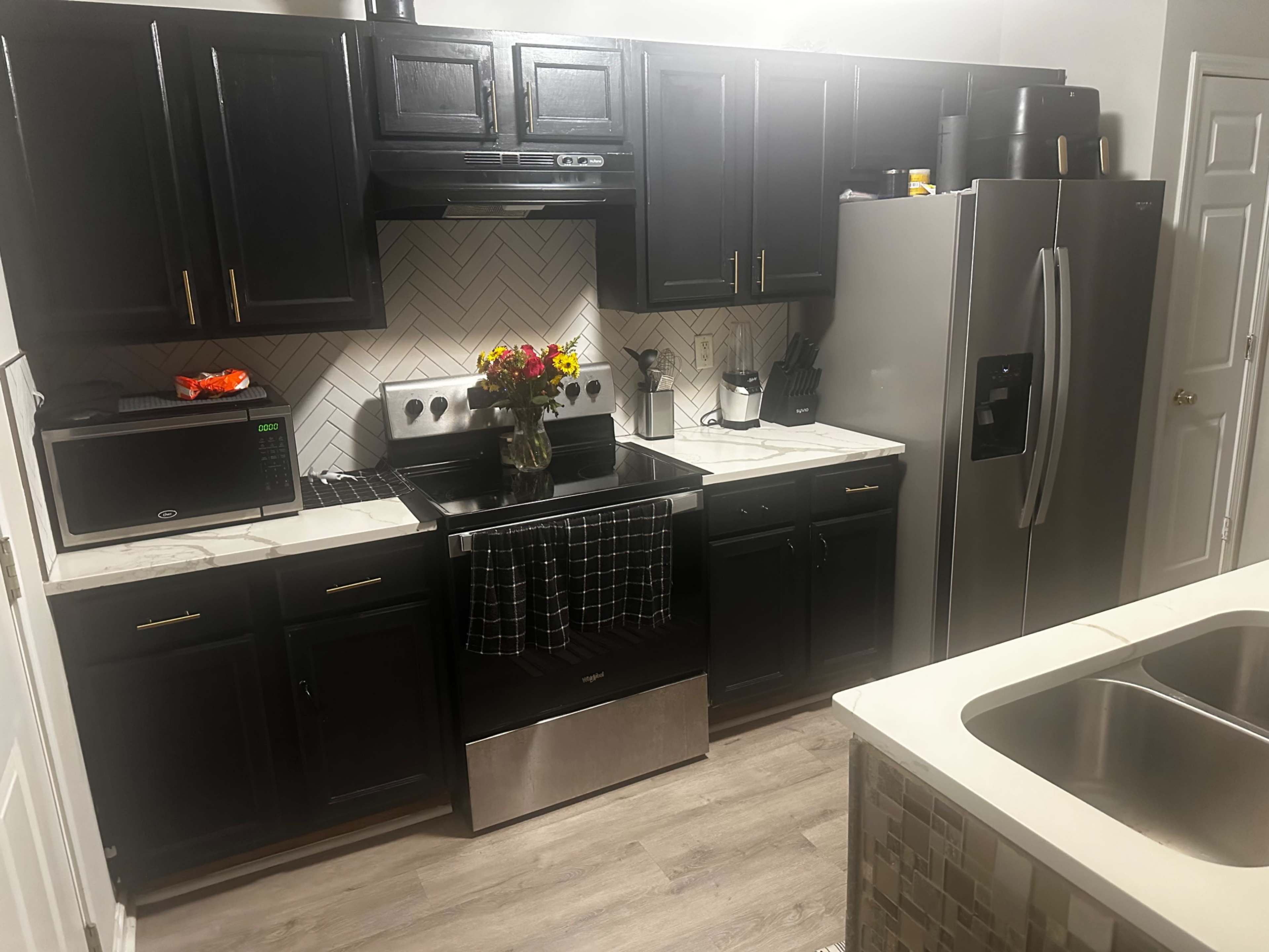 The image shows a modern kitchen featuring dark cabinetry, stainless steel appliances, a marble countertop, and a bouquet of flowers on the counter.